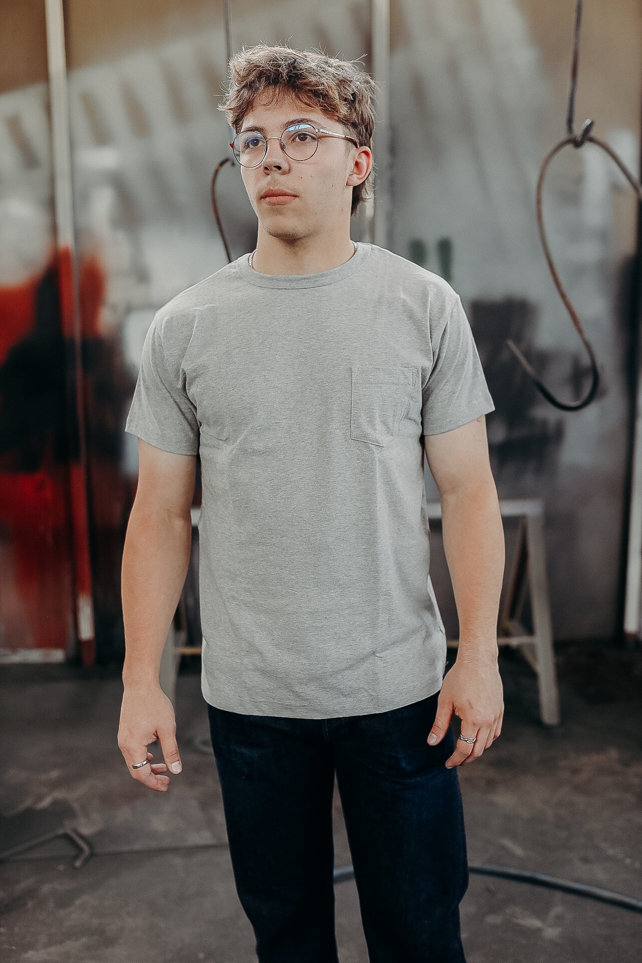 Young man wearing gray pocket t-shirt and glasses standing in industrial workshop setting