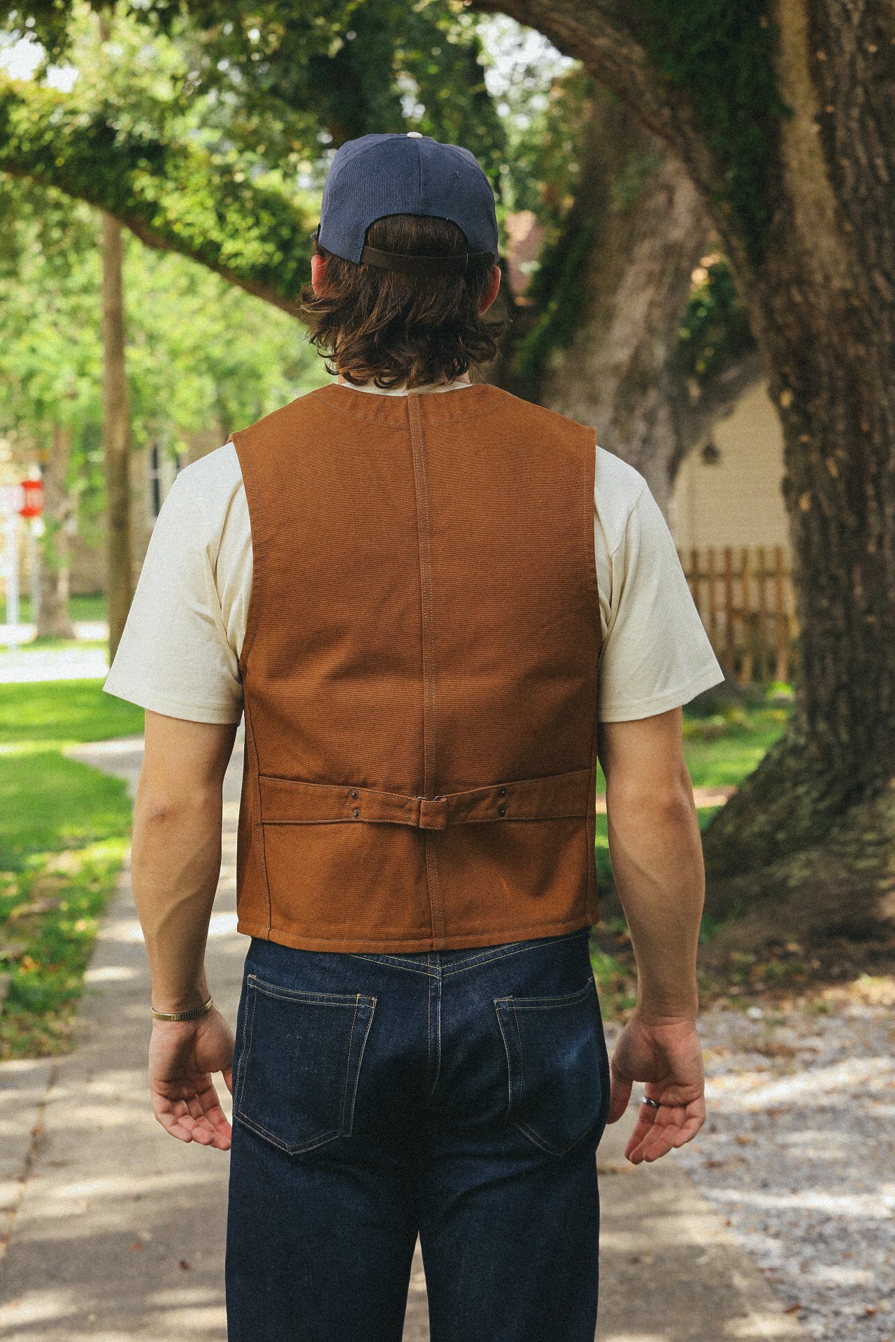 Man wearing brown work vest over cream t-shirt and blue jeans standing outdoors near trees