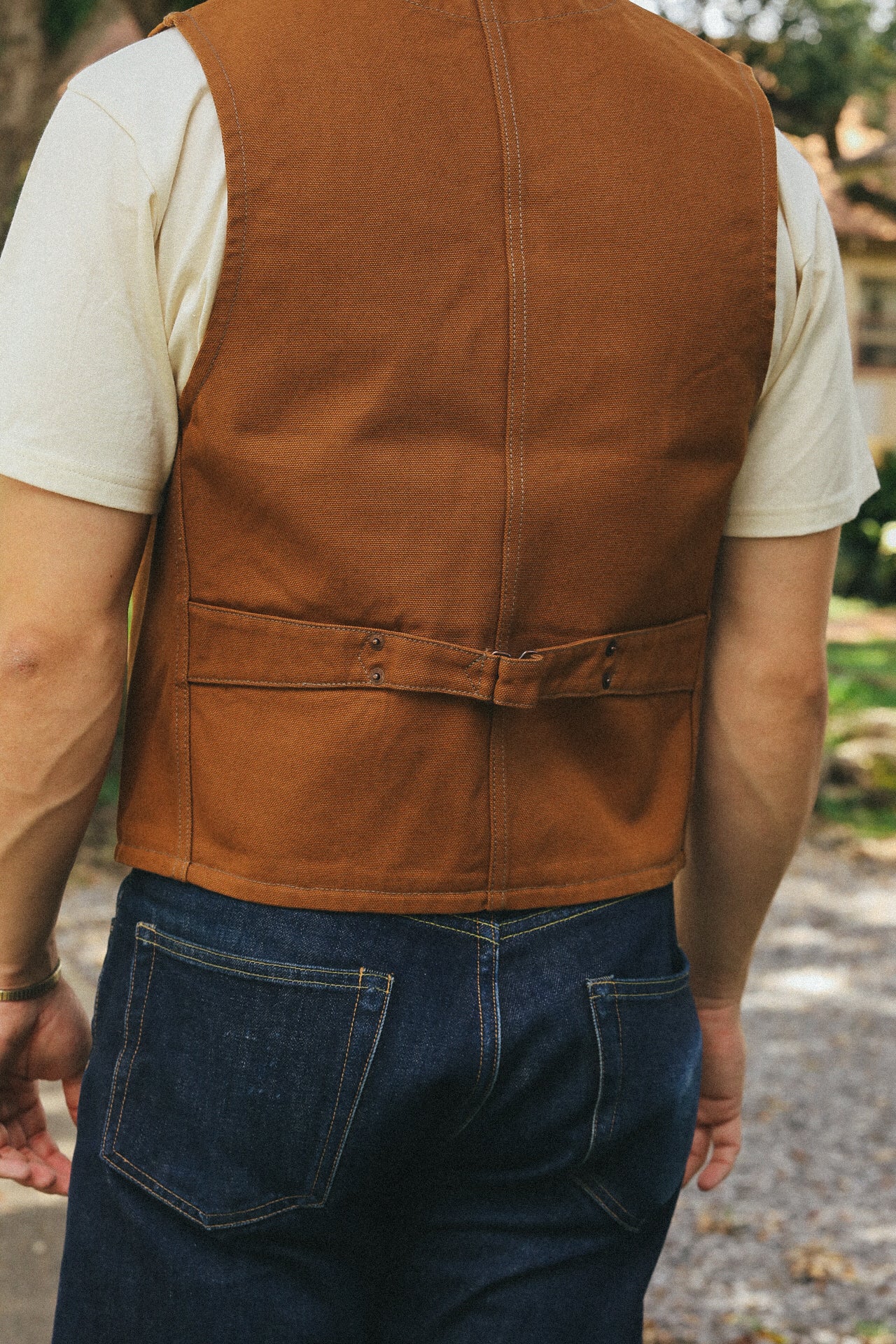 Back view of a person wearing a brown canvas work vest over a white t-shirt with dark denim jeans, outdoors