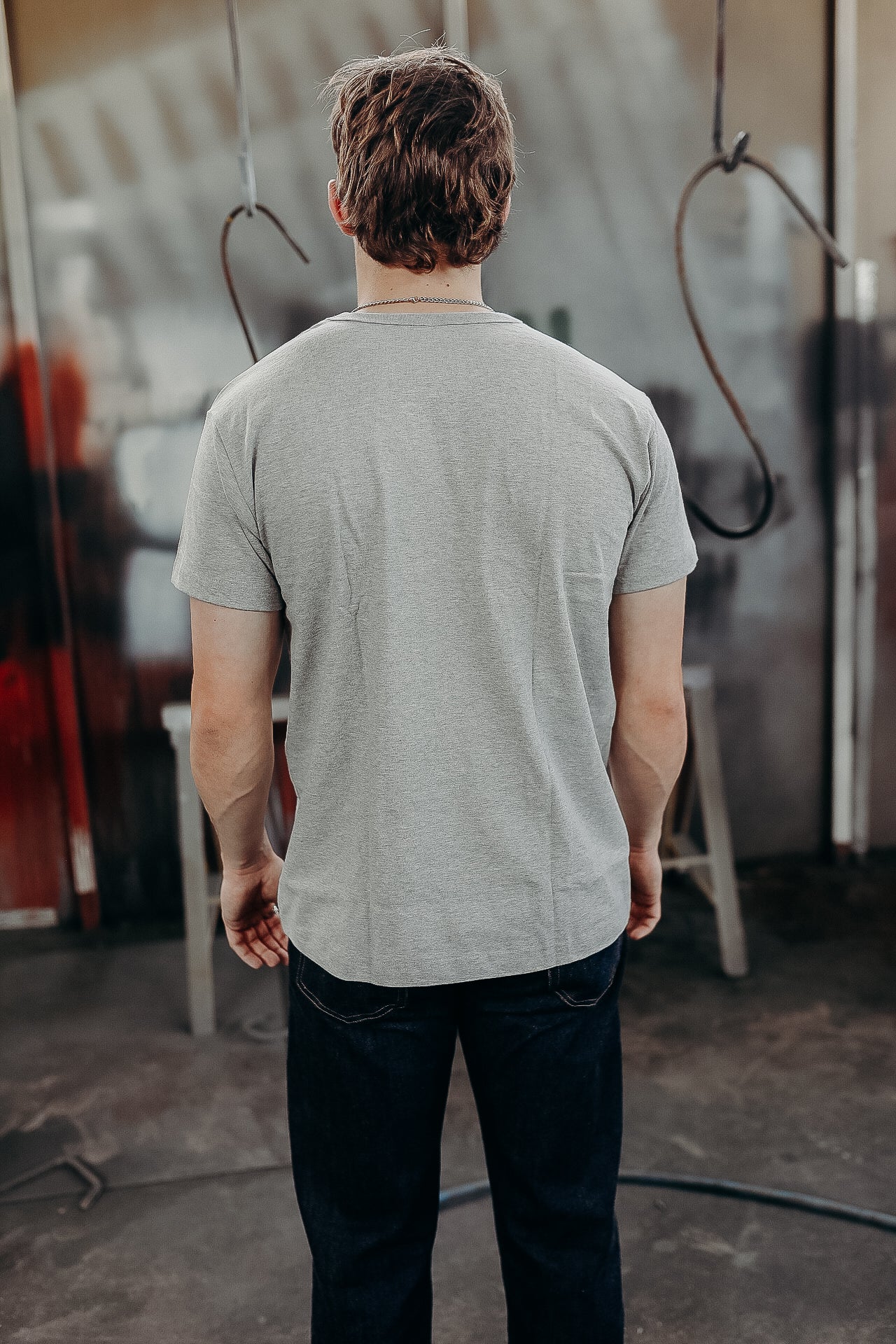 Man wearing plain gray t-shirt and dark jeans standing in industrial workshop interior with metal hooks