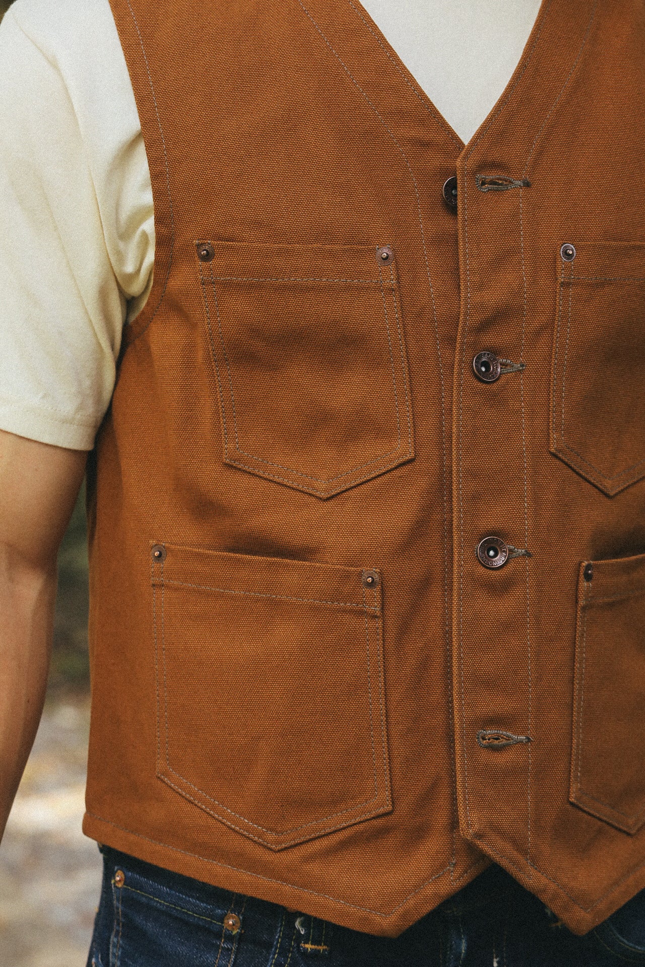 close-up of a person wearing a rugged brown canvas work vest with multiple pockets over a white t-shirt