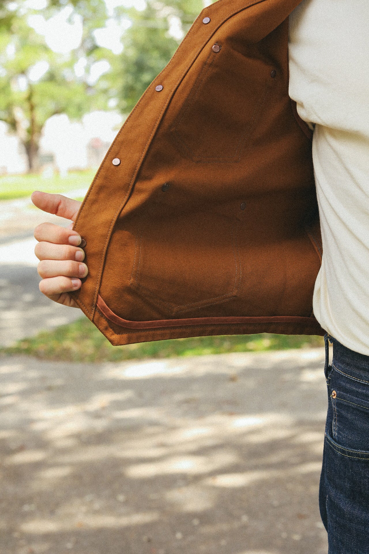 Close-up of a person wearing a white shirt and dark jeans holding open a brown rugged work jacket outdoors