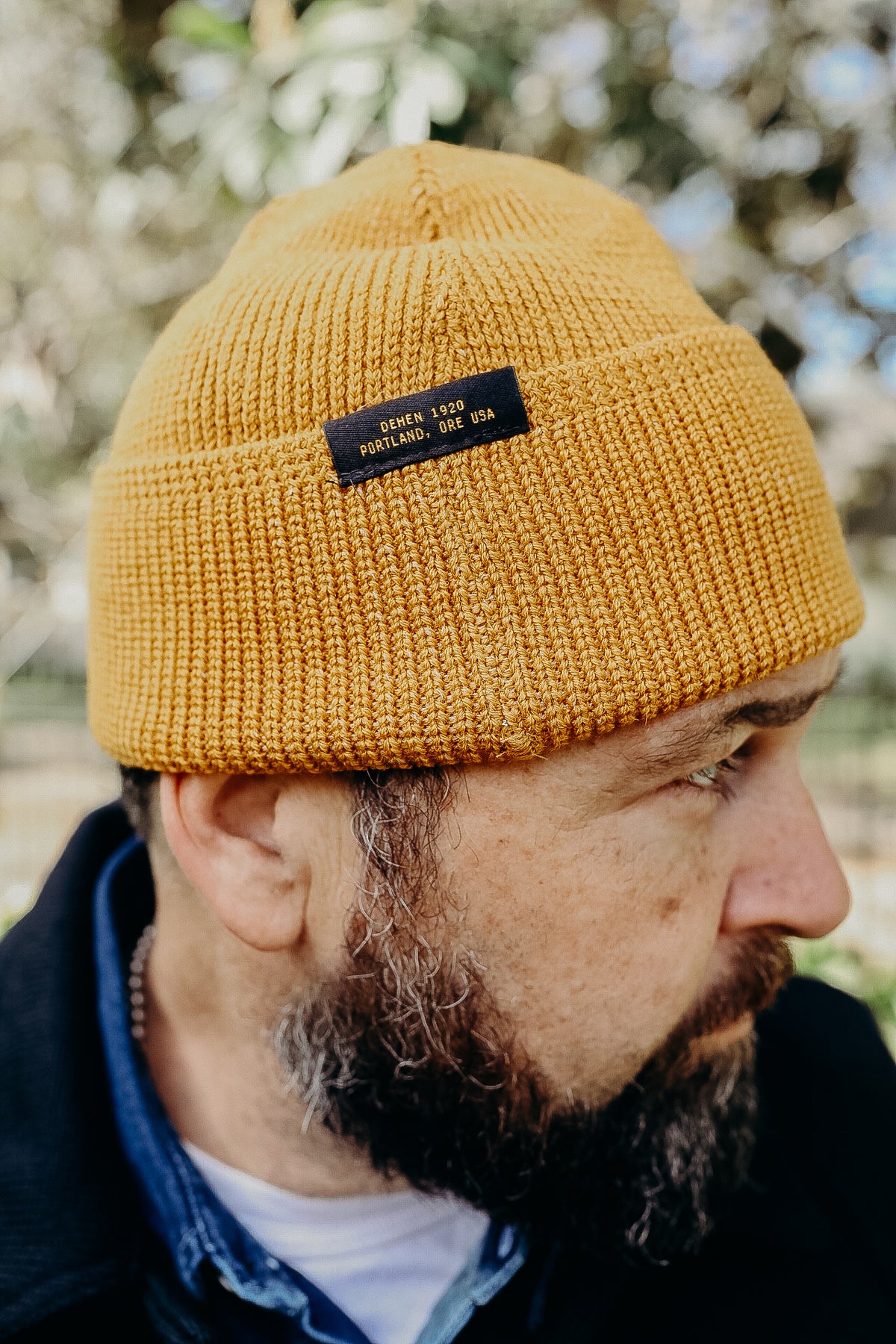 Side profile of a man wearing a mustard yellow knit beanie with a black label reading Dehen 1920 Portland, ORE USA