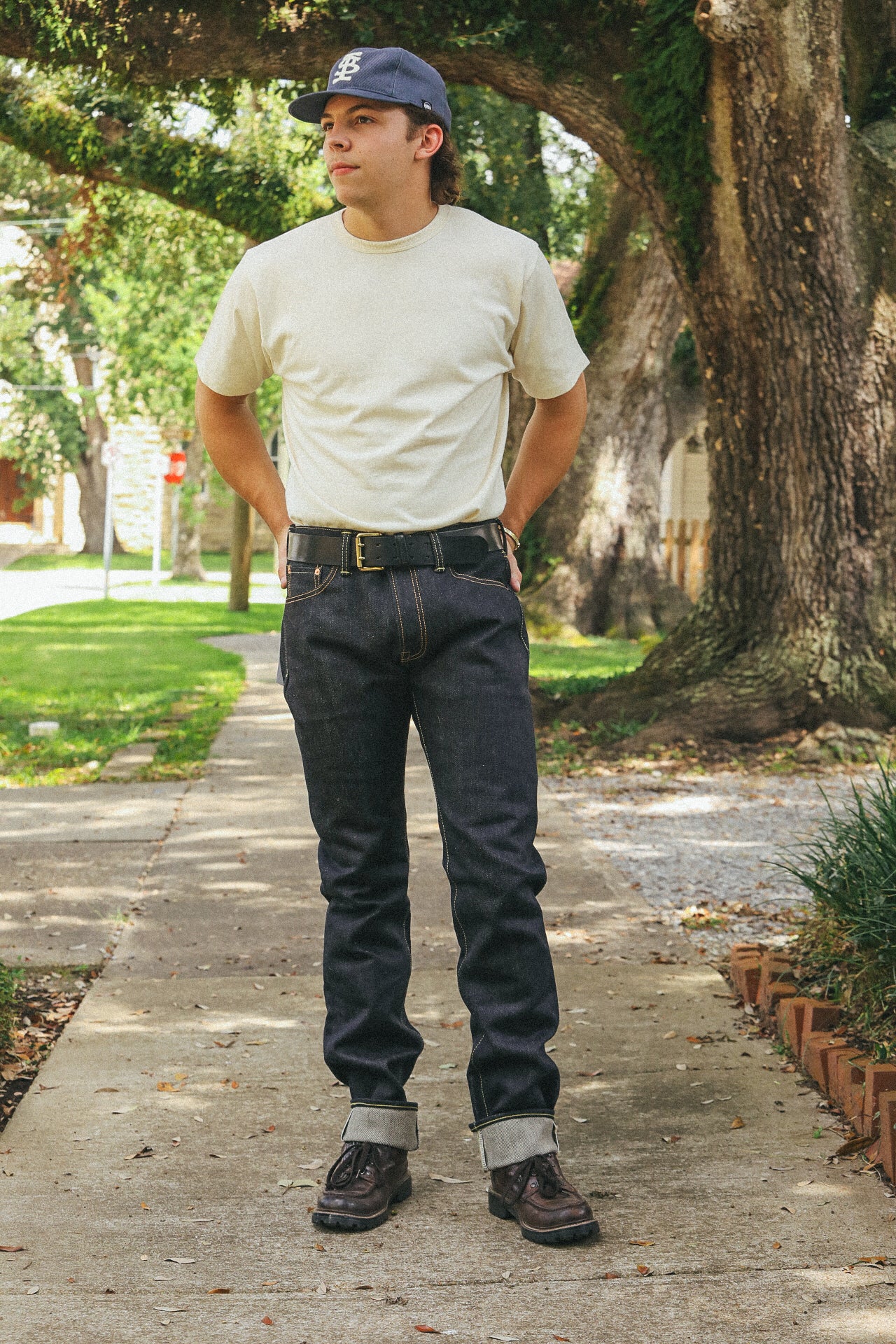 Young man wearing cream t-shirt, dark selvedge jeans, leather belt, and brown boots standing on sidewalk under large tree