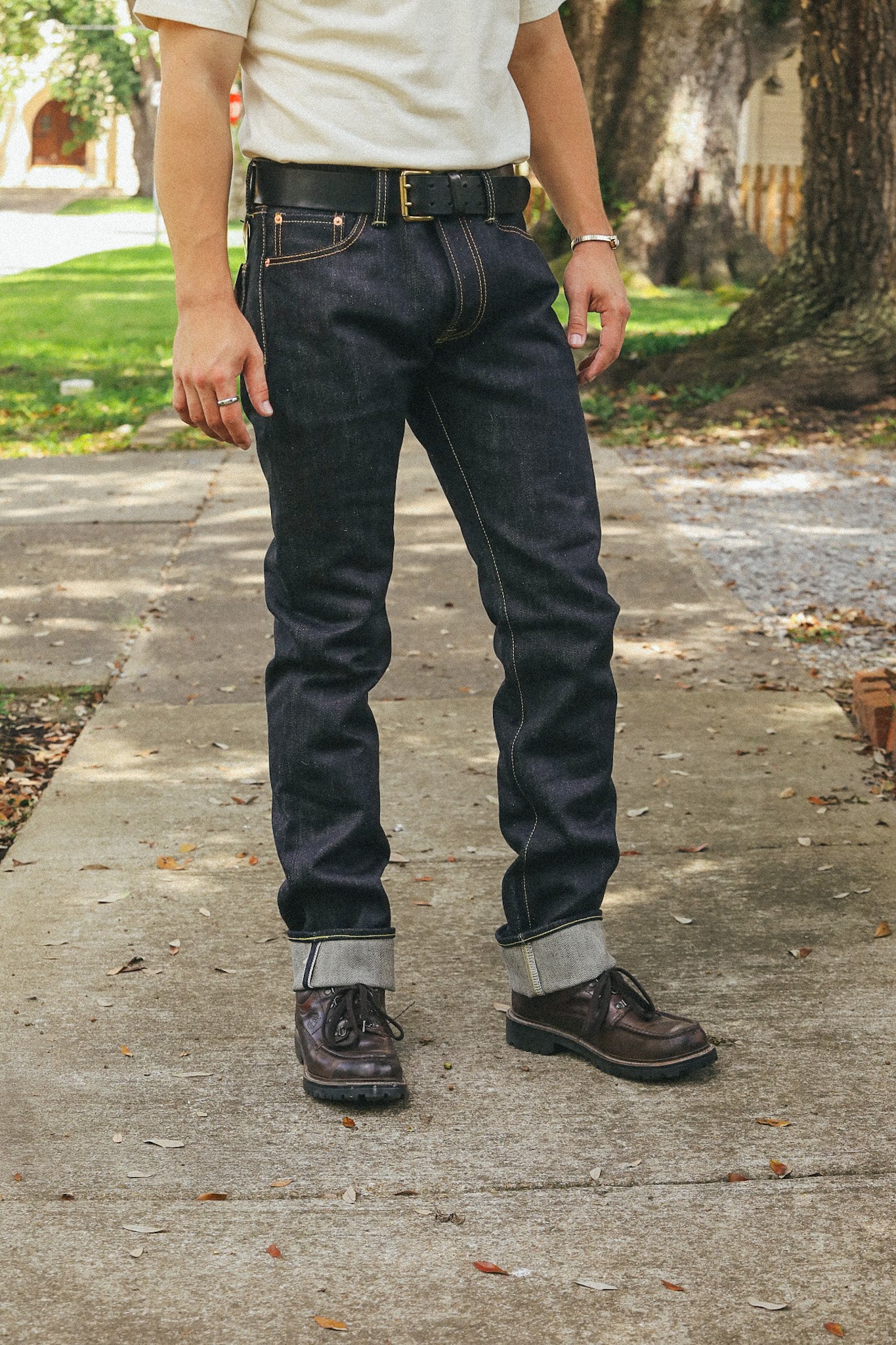 Man wearing dark raw denim jeans with rolled cuffs, black belt, and brown leather boots on sidewalk
