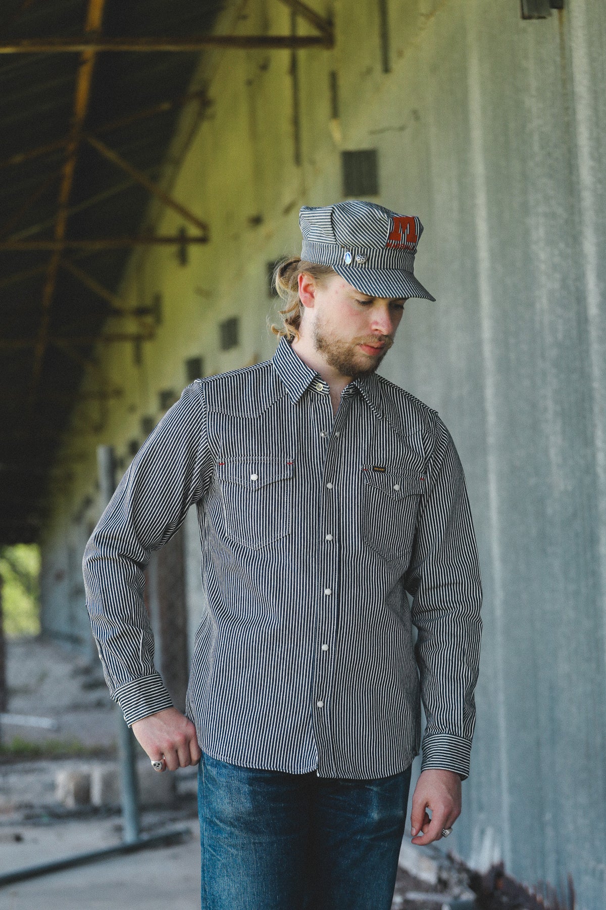 A man wearing a gray and black vertically striped button-down shirt and a matching gray cap with a red logo, standing in an industrial warehouse setting.