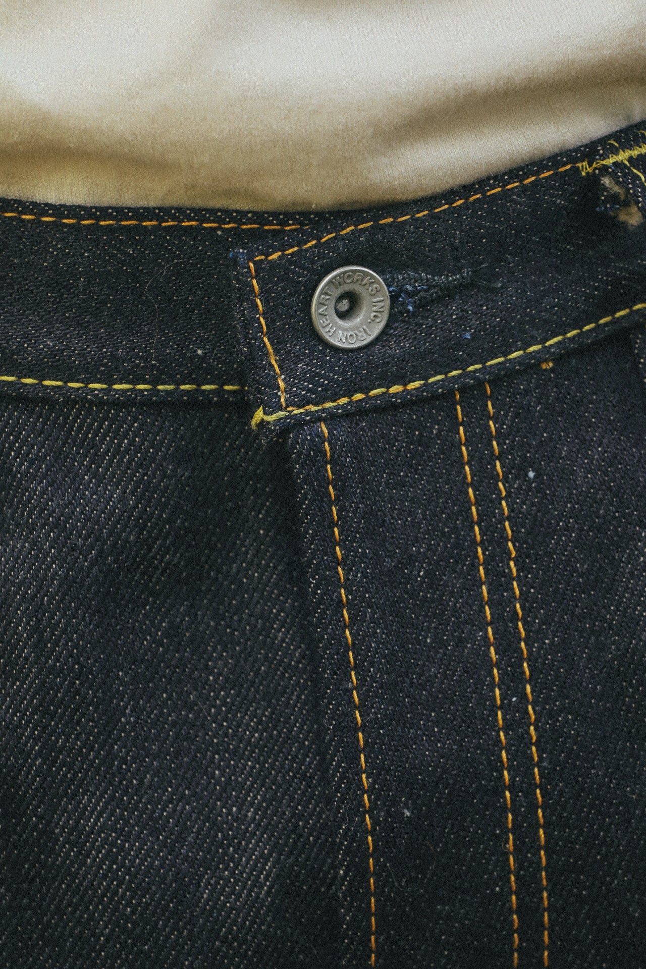 Close-up of dark denim jeans waistband with yellow stitching and a metal button engraved with Iron Heart Works Inc.