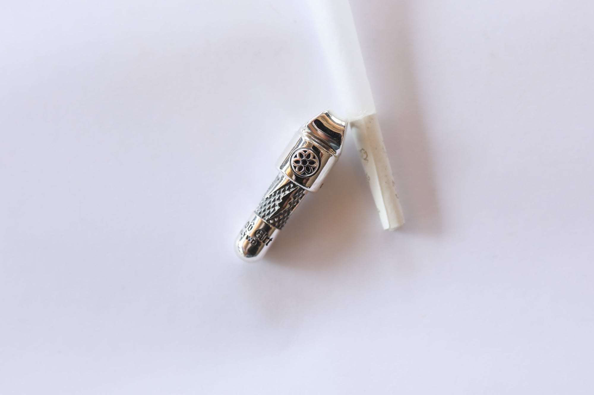 Close-up of a silver metal pipe with intricate engravings next to a broken white cigarette on a white background