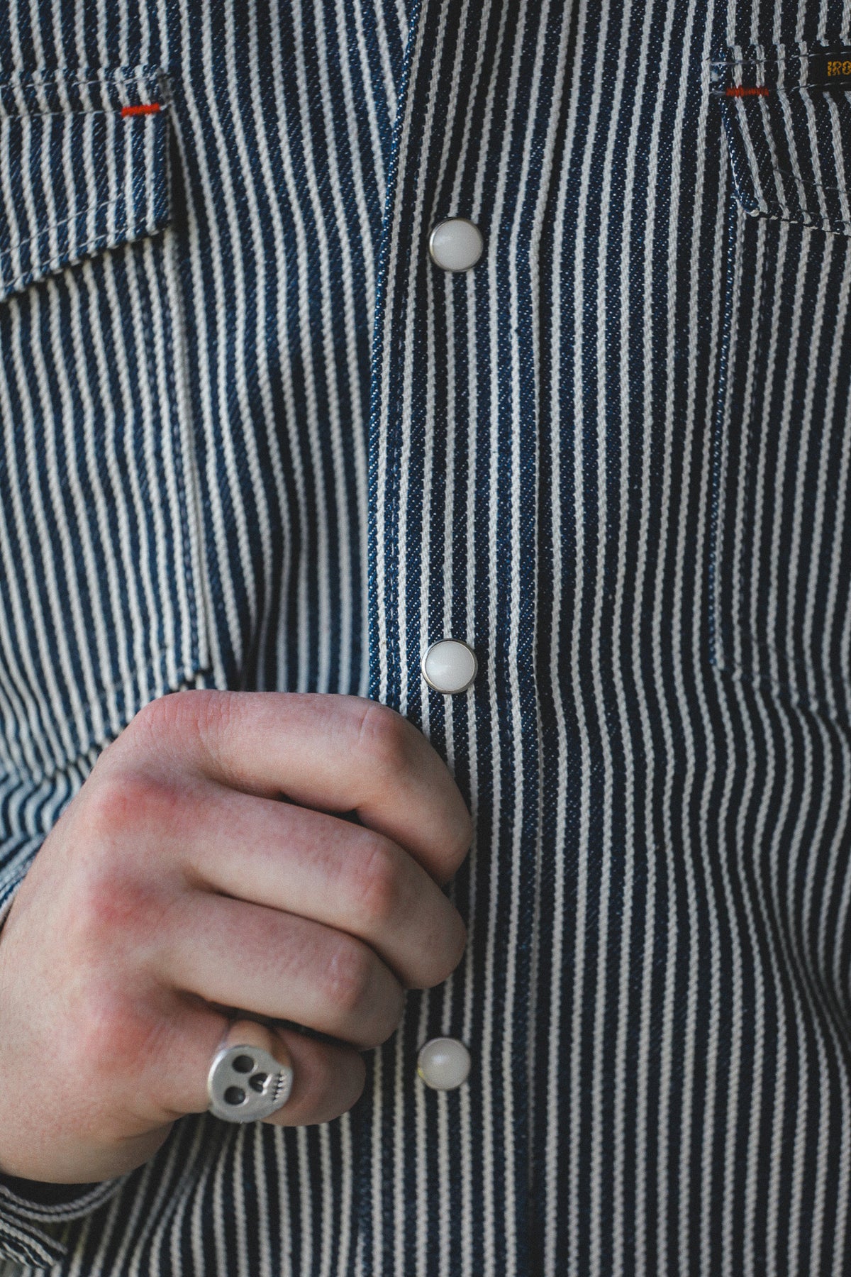 Close-up of a person wearing a navy and white vertically striped button-down shirt, with a hand buttoning the front and a skull ring visible on the finger.