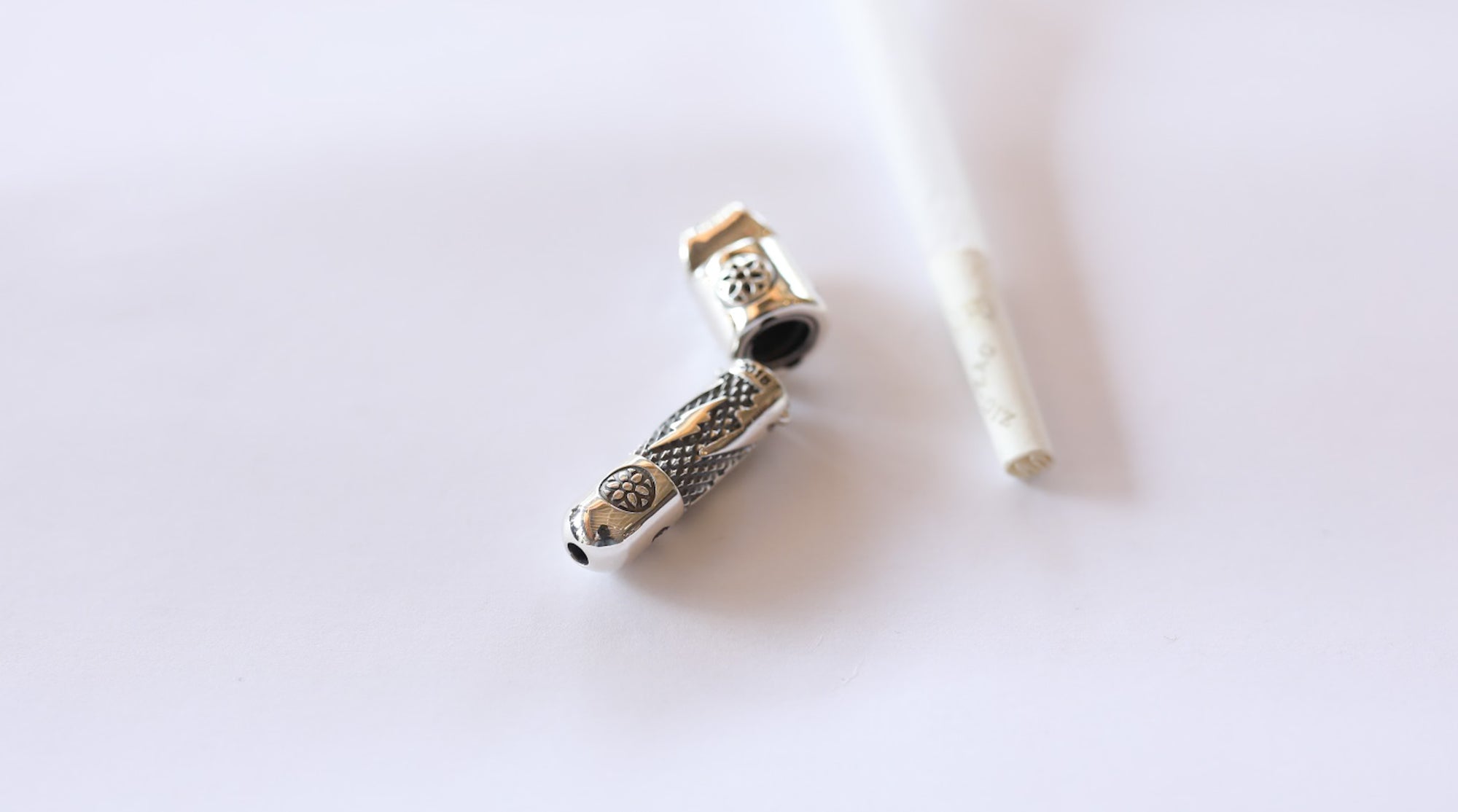 open silver metal cigarette case with intricate designs beside a white cigarette on white background