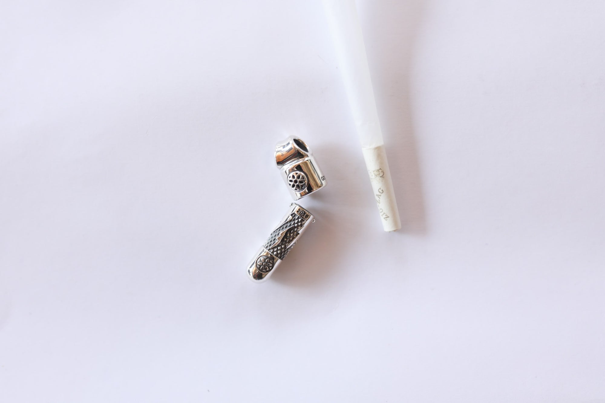 open silver metal pill case with engraved designs beside a cigarette on a white background