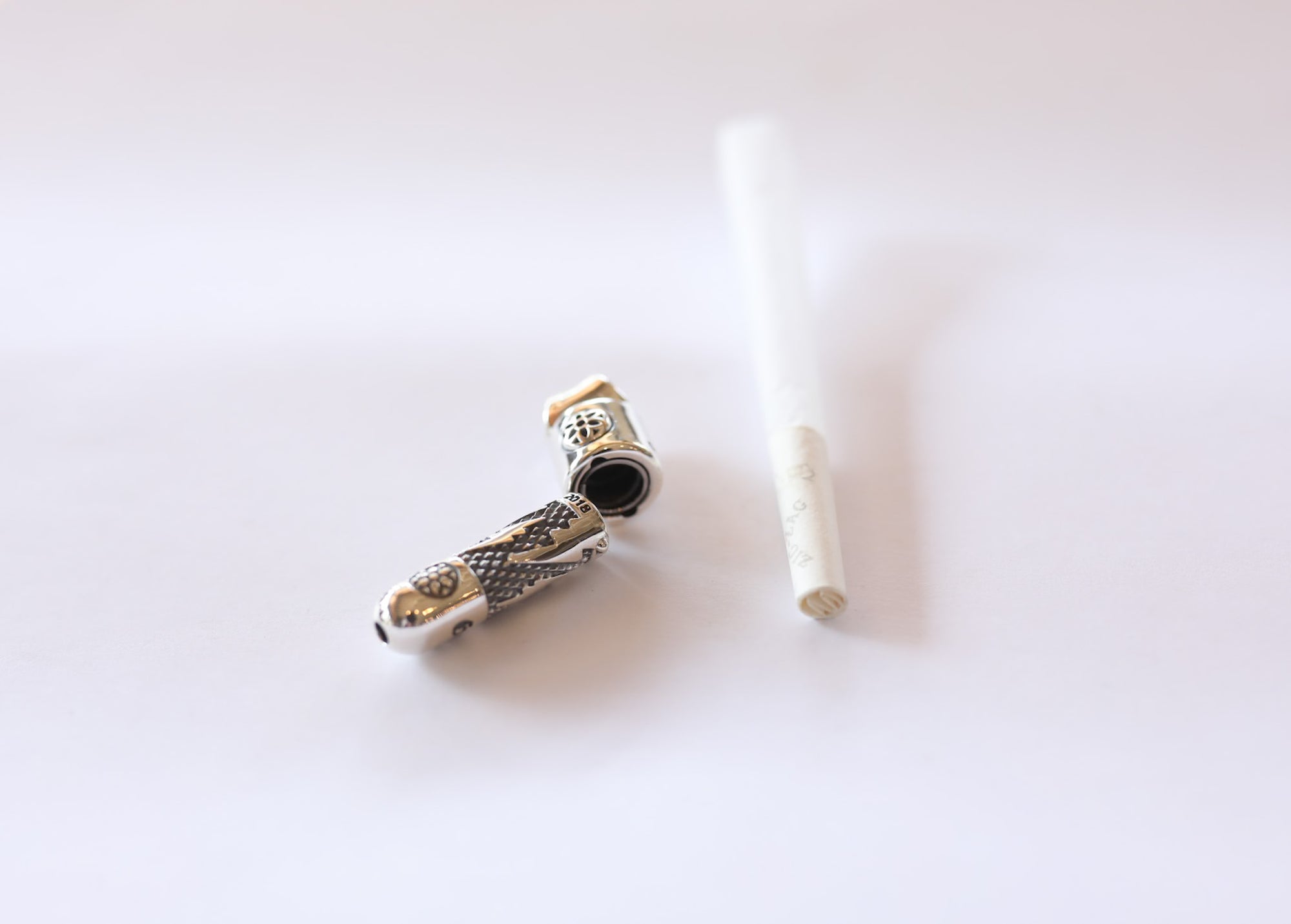 Small metal smoking pipe with dragon design and white rolled cigarette on white surface