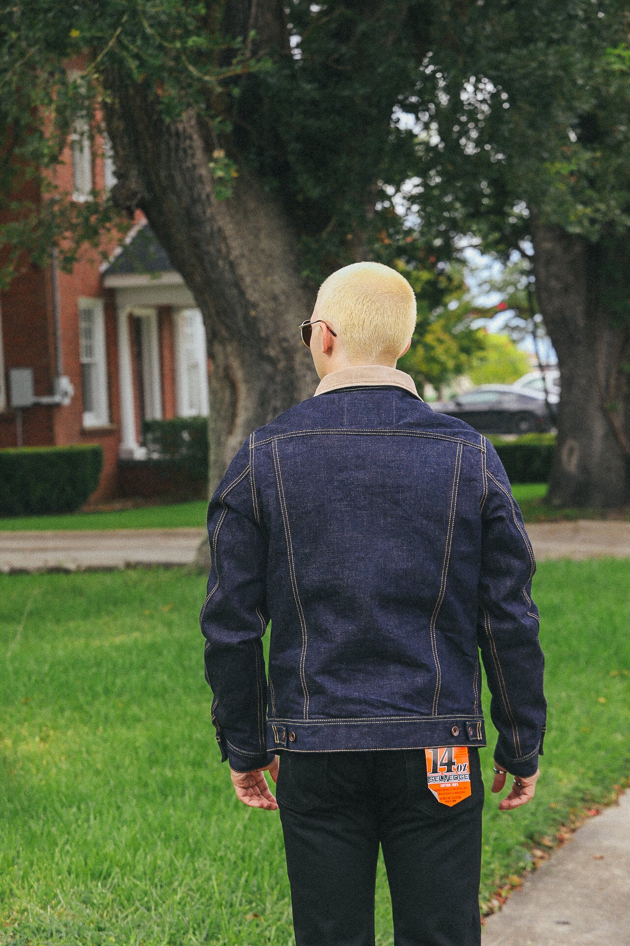 Man with platinum blonde hair wearing dark blue denim jacket and black pants outdoors on grassy walkway