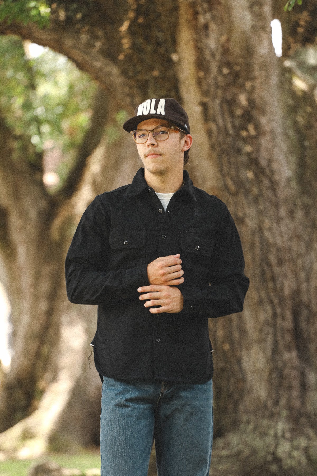Person wearing a black jacket and cap with 'Bola' branding, standing in front of a tree.