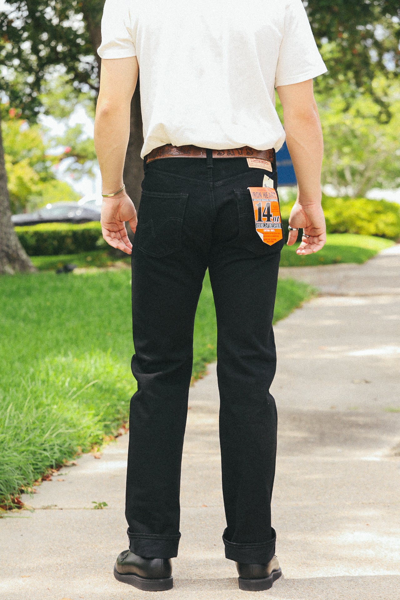Man wearing black Iron Heart 14 oz selvedge denim jeans with a white shirt, leather belt, and black shoes on a sidewalk