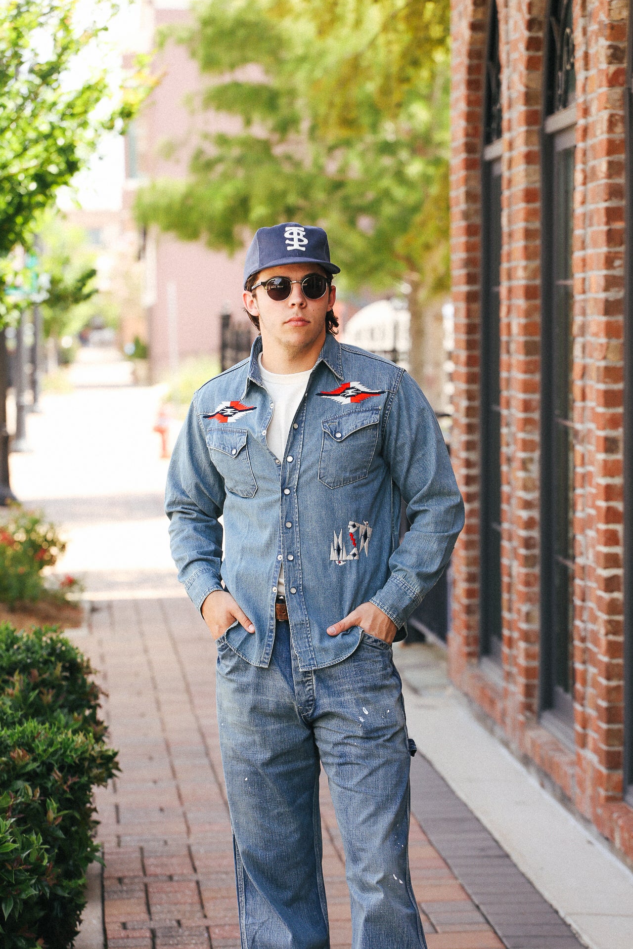 Man wearing embroidered denim western shirt and jeans with sunglasses and a navy cap on city sidewalk