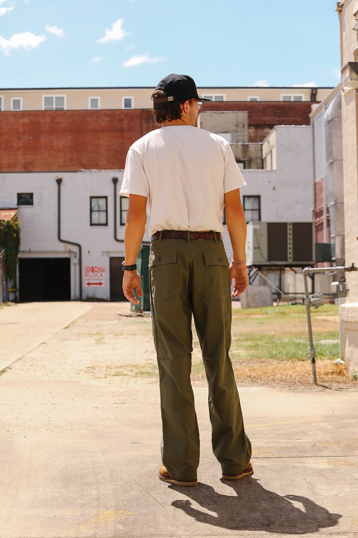A man viewed from behind wearing olive green wide-leg work pants, a white t-shirt, and a black baseball cap, standing on a concrete walkway in an outdoor industrial setting.
