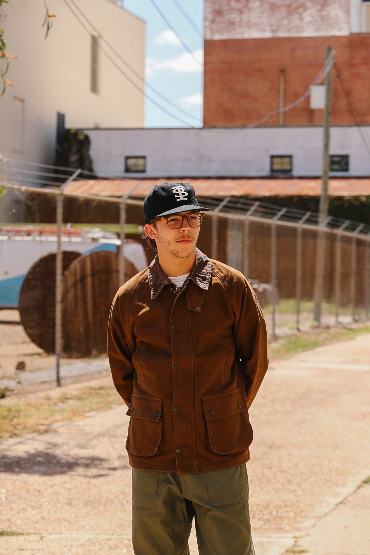 A man wearing a brown corduroy jacket, olive green pants, a black baseball cap, and glasses stands on an outdoor paved walkway with industrial buildings and a chain-link fence in the background.