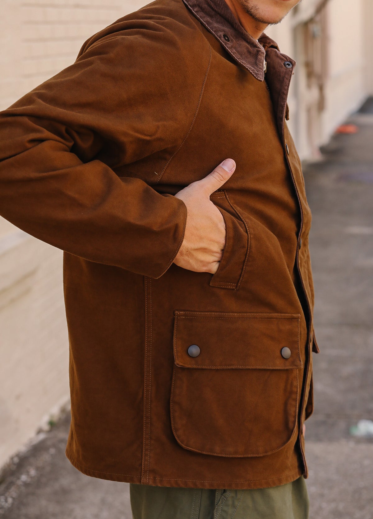 A man wearing a brown corduroy jacket with a snap-button front pocket, shown from the chest down in an outdoor alley setting.