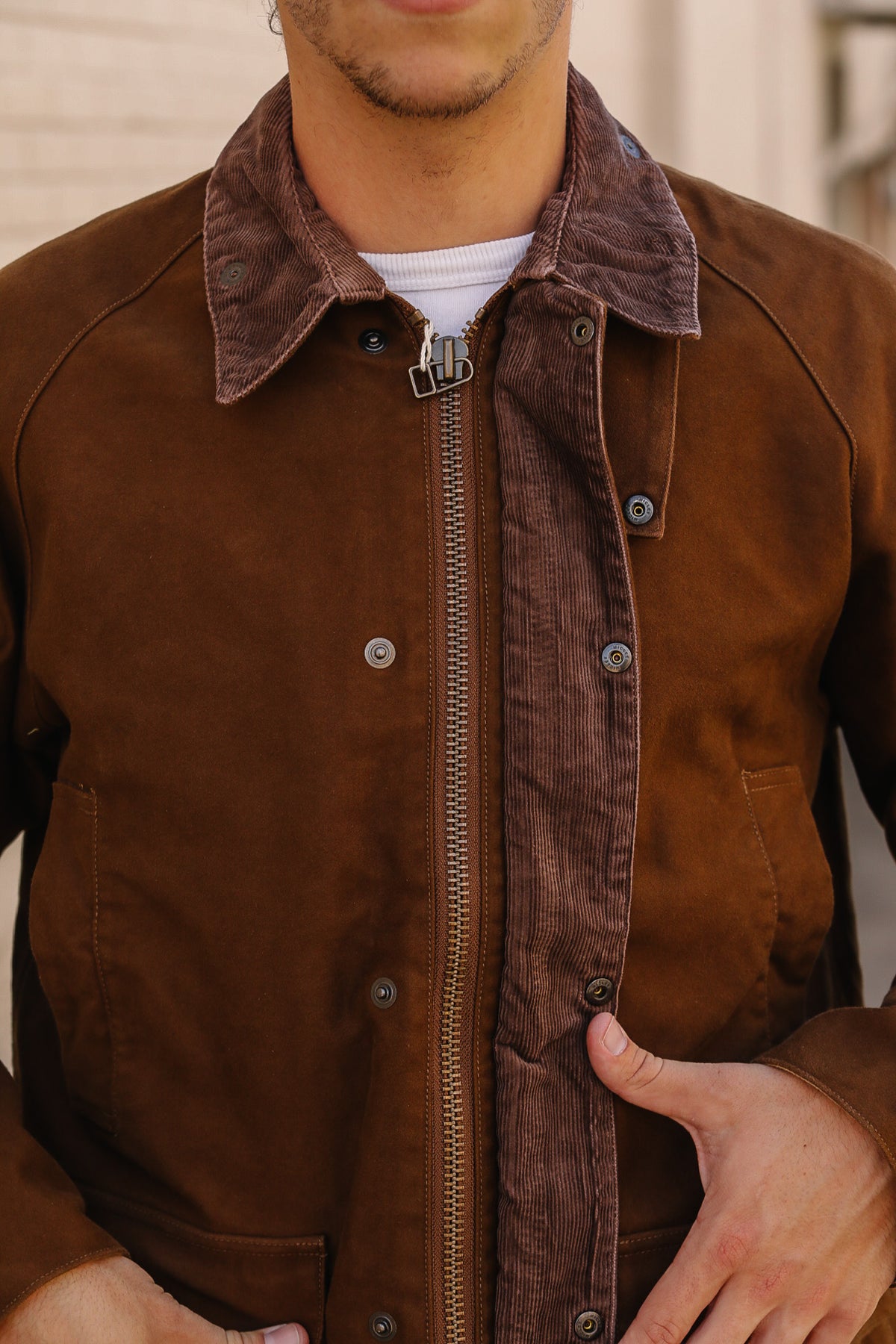 A close-up of a person wearing a brown jacket with a brown corduroy collar and front zipper, layered over a white t-shirt.
