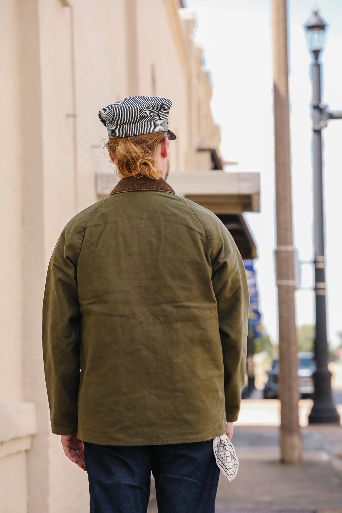 A person viewed from behind wearing an olive green canvas utility jacket with a brown collar, a gray striped flat cap, and dark blue jeans, standing on a city sidewalk.