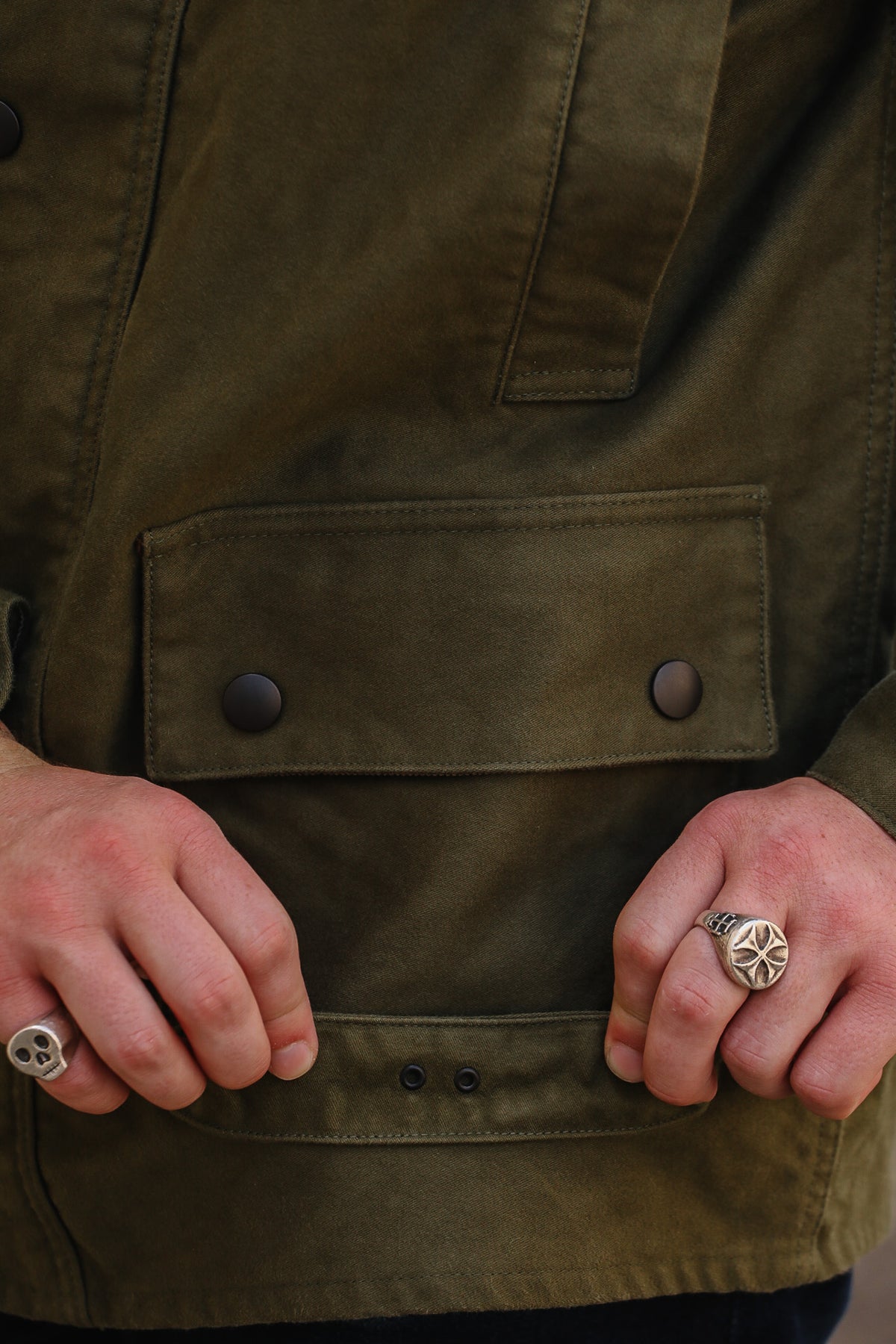 Close-up of a person wearing an olive green waxed canvas utility jacket, showing the front flap pocket with snap buttons and the hands holding the snap tab open.
