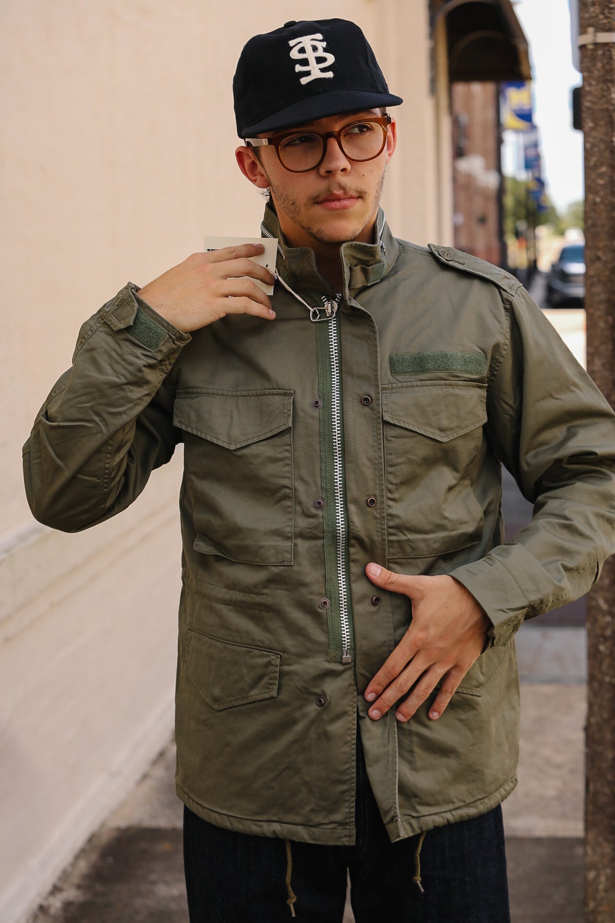 A man wearing an olive green military-style field jacket with a front zipper, chest pockets, and a black baseball cap with a white logo, standing outdoors on a city street.