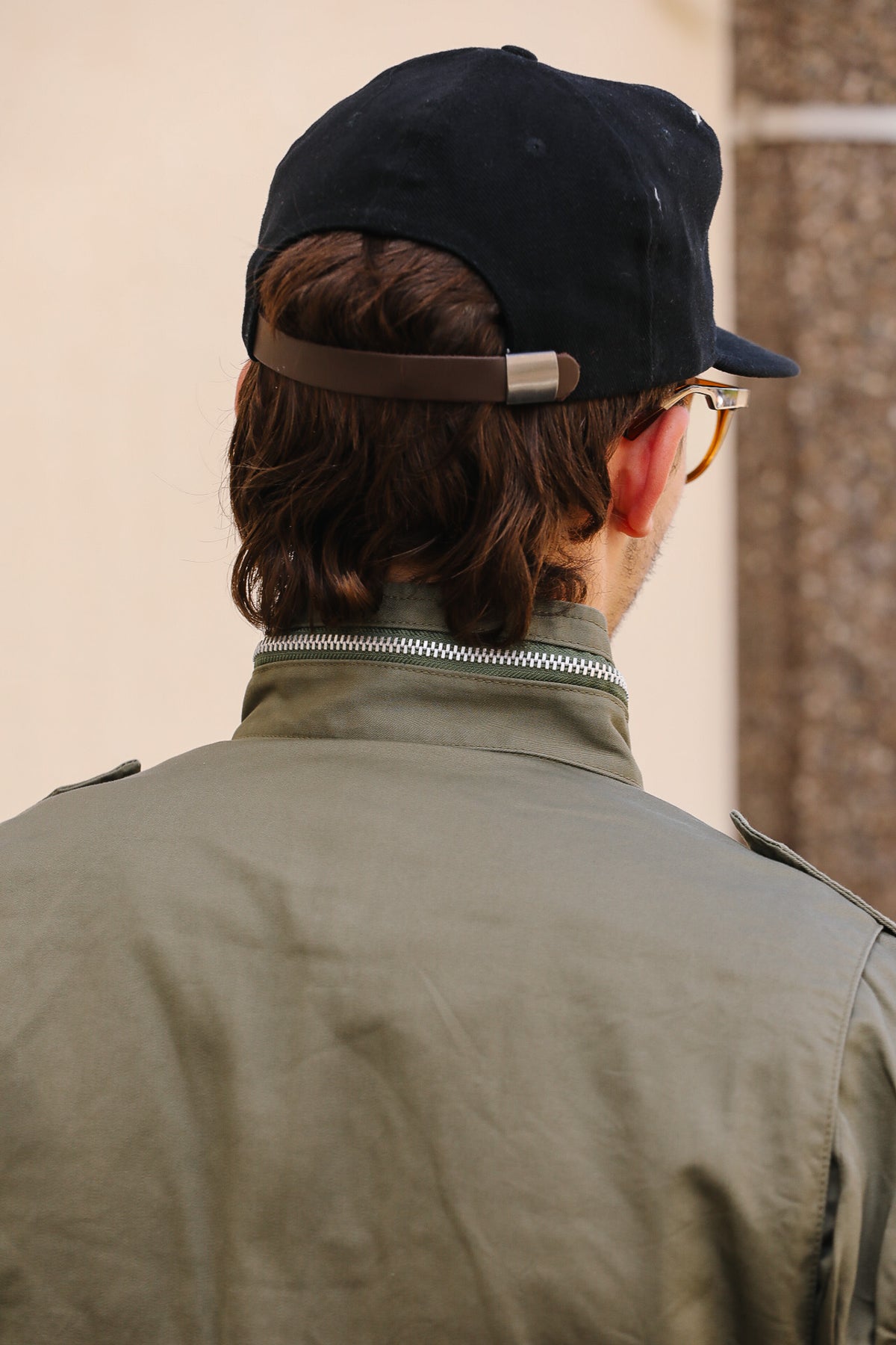 Back view of a person wearing a dark navy baseball cap with a brown adjustable strap, glasses, and an olive green jacket.