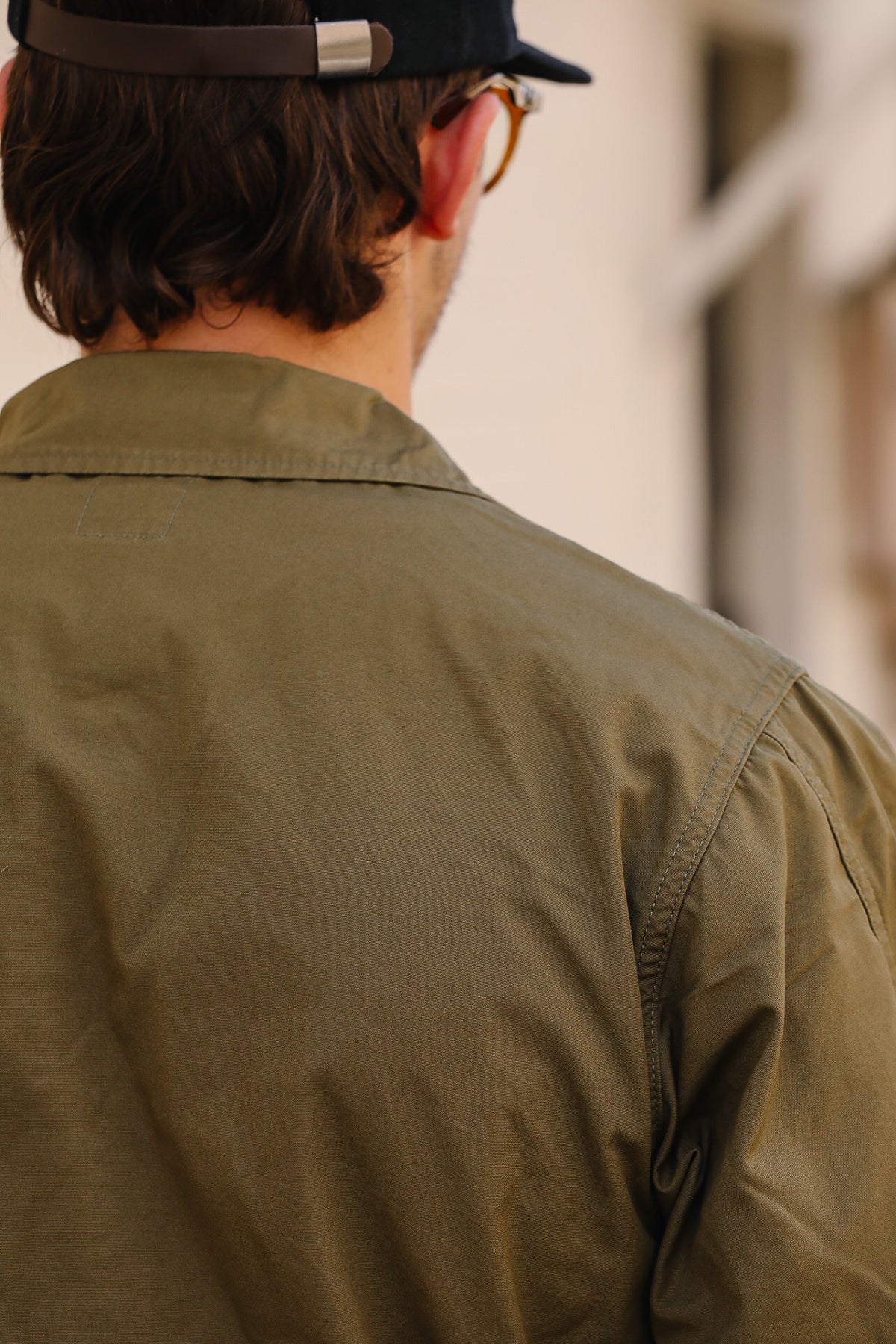 A man viewed from behind wearing an olive green utility jacket, a black cap, and glasses, set against a softly blurred beige background.