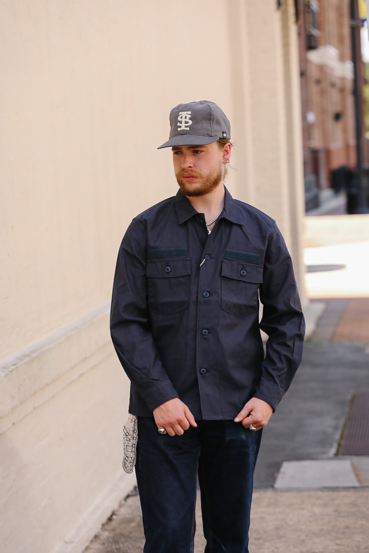 A man wearing a dark navy blue button-up work shirt, dark jeans, and a gray baseball cap with a white 'BF' logo, standing on a city sidewalk next to a beige wall.