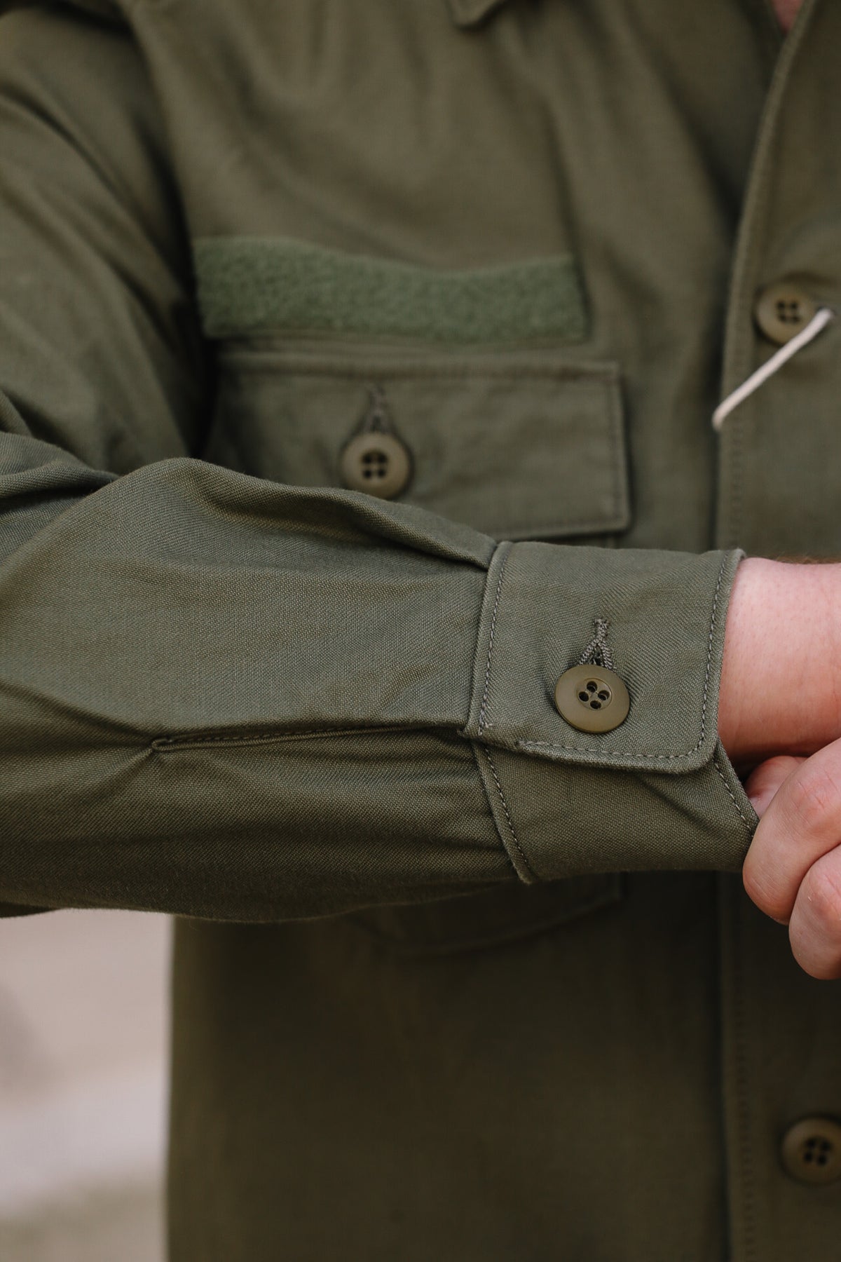 Close-up of a person wearing an olive green utility shirt jacket, focusing on the buttoned cuff and chest pocket details.