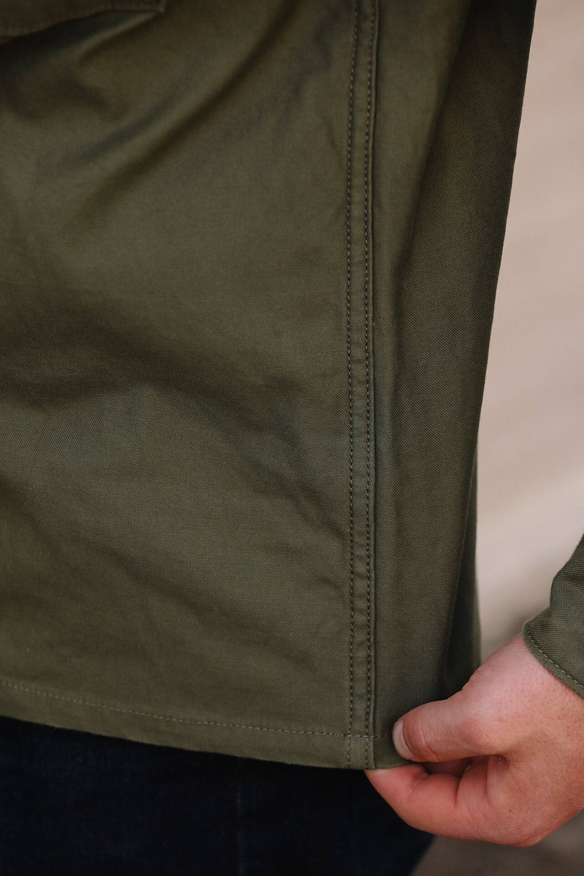 Close-up of an olive green jacket or overshirt being held at the hem by a person's hand, showing the fabric texture and stitching against a neutral background.