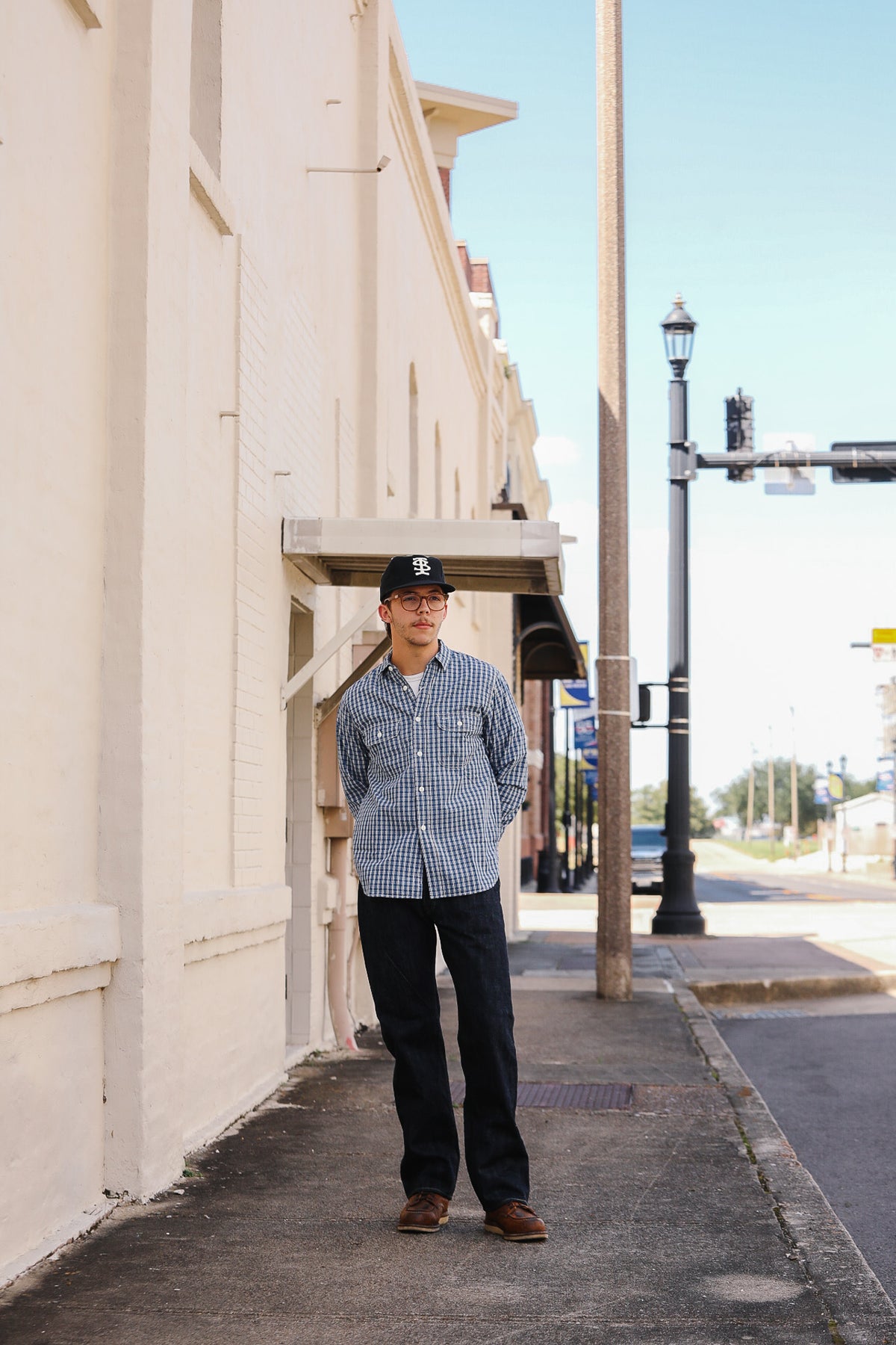 A man wearing a blue and white checkered button-down shirt, dark pants, and a baseball cap stands on a city sidewalk next to a beige building.