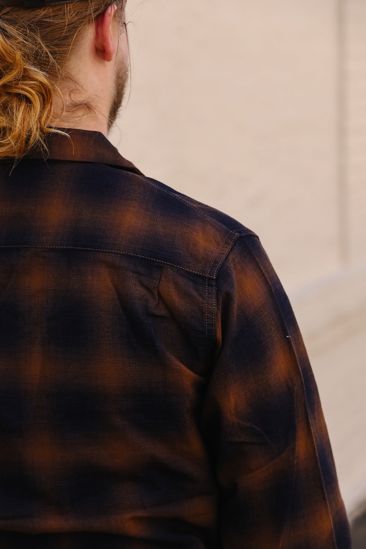 A close-up back view of a person wearing a dark brown and black plaid flannel shirt, with light beige wall in the background.