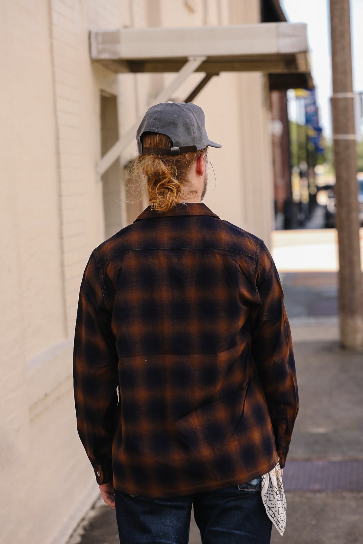 A person viewed from behind wearing a dark brown and black plaid flannel shirt, a gray baseball cap, and dark jeans, standing on a city sidewalk next to a beige brick building.