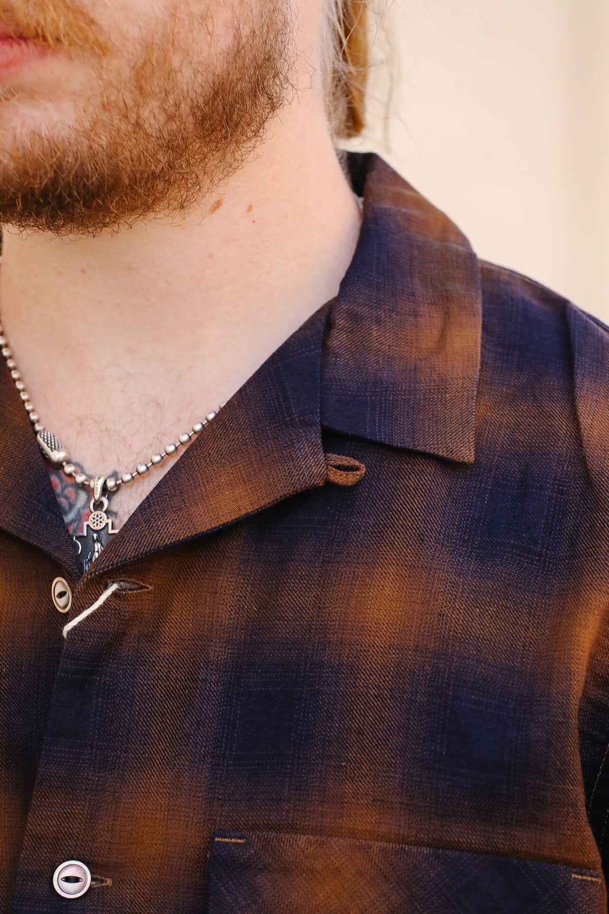 Close-up of a man wearing a brown and navy plaid button-down shirt with a collar and chest pocket, accessorized with a silver chain necklace.