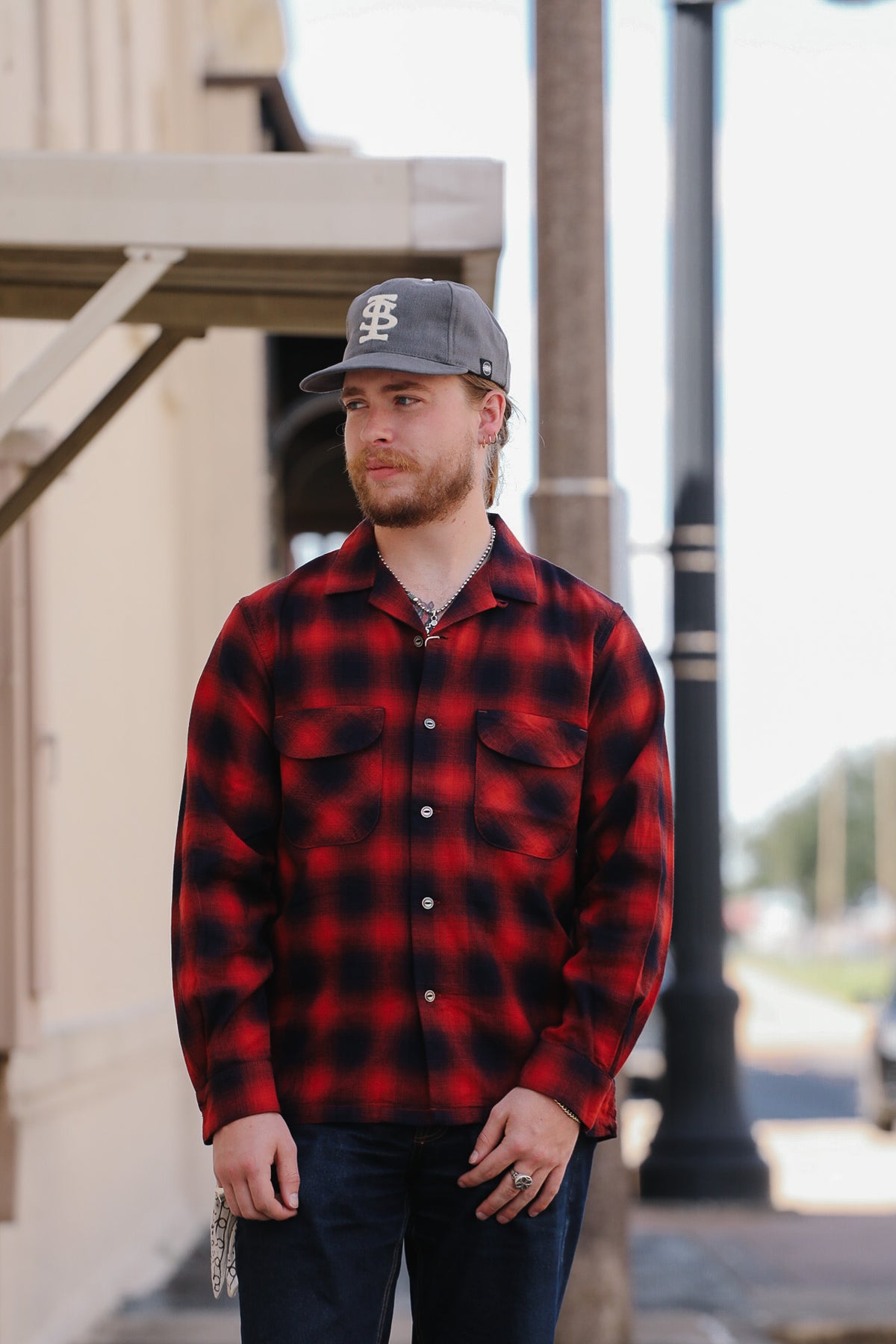 A man wearing a red and black buffalo plaid flannel shirt, dark jeans, and a gray baseball cap with a white logo, standing outdoors on a city street.