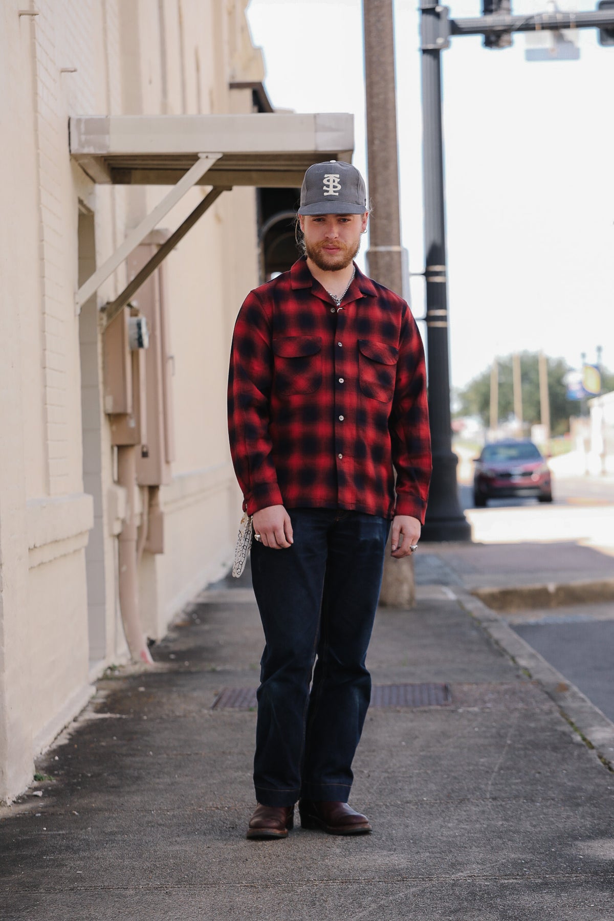 A man standing on a city sidewalk wearing a red and black buffalo plaid flannel shirt, dark jeans, brown boots, and a gray baseball cap with a white logo.