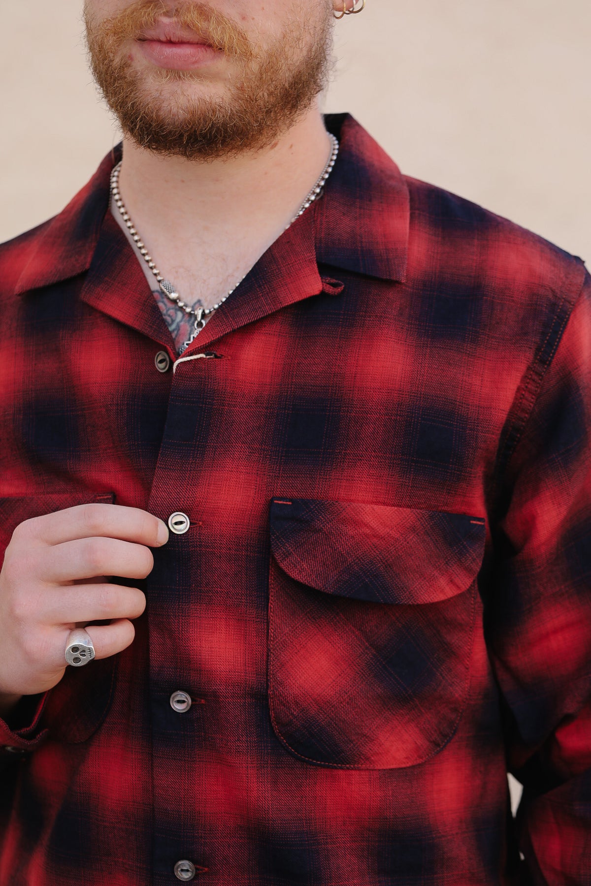 A close-up of a man wearing a red and black plaid button-down shirt with a chest pocket, layered over a silver chain necklace.