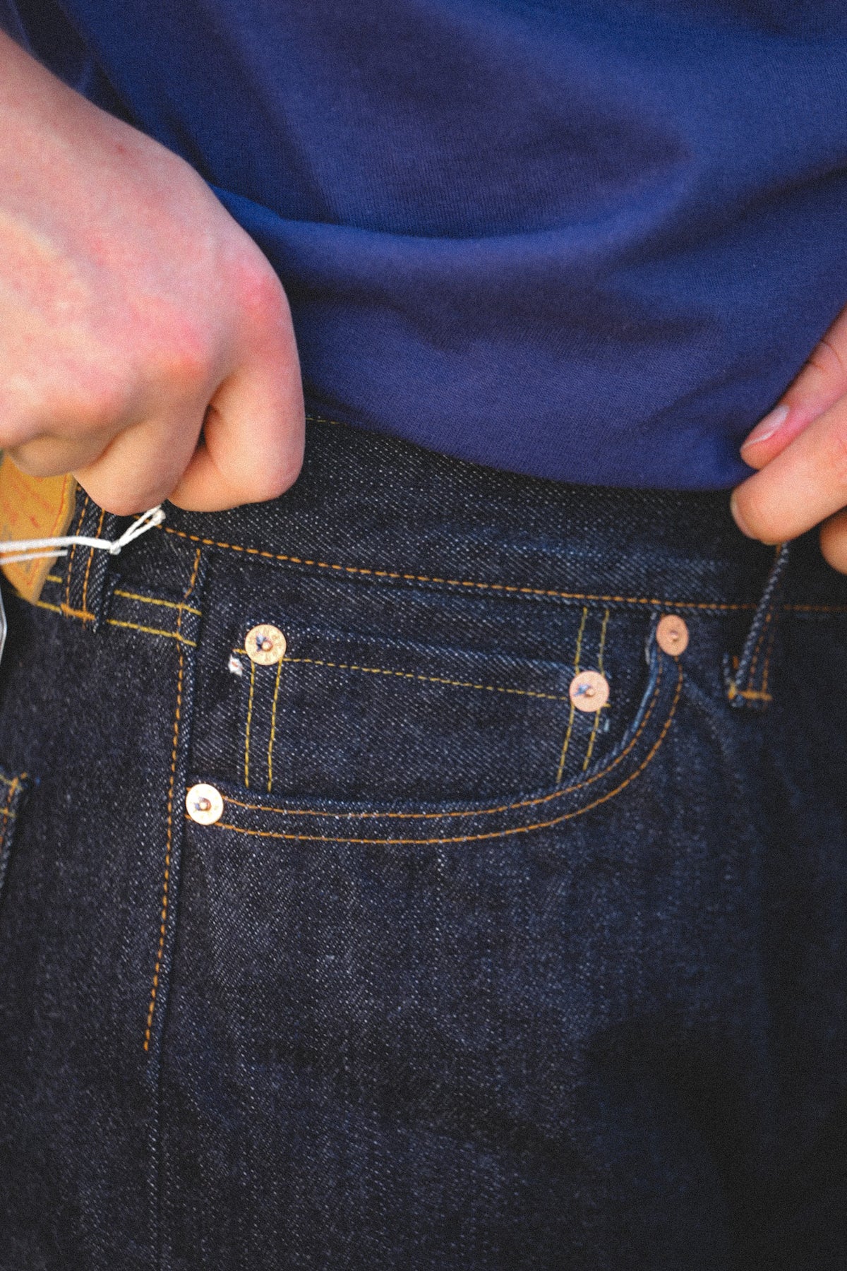 Close-up of a person's hands adjusting the waistband of dark blue denim jeans, with a navy blue t-shirt visible above.