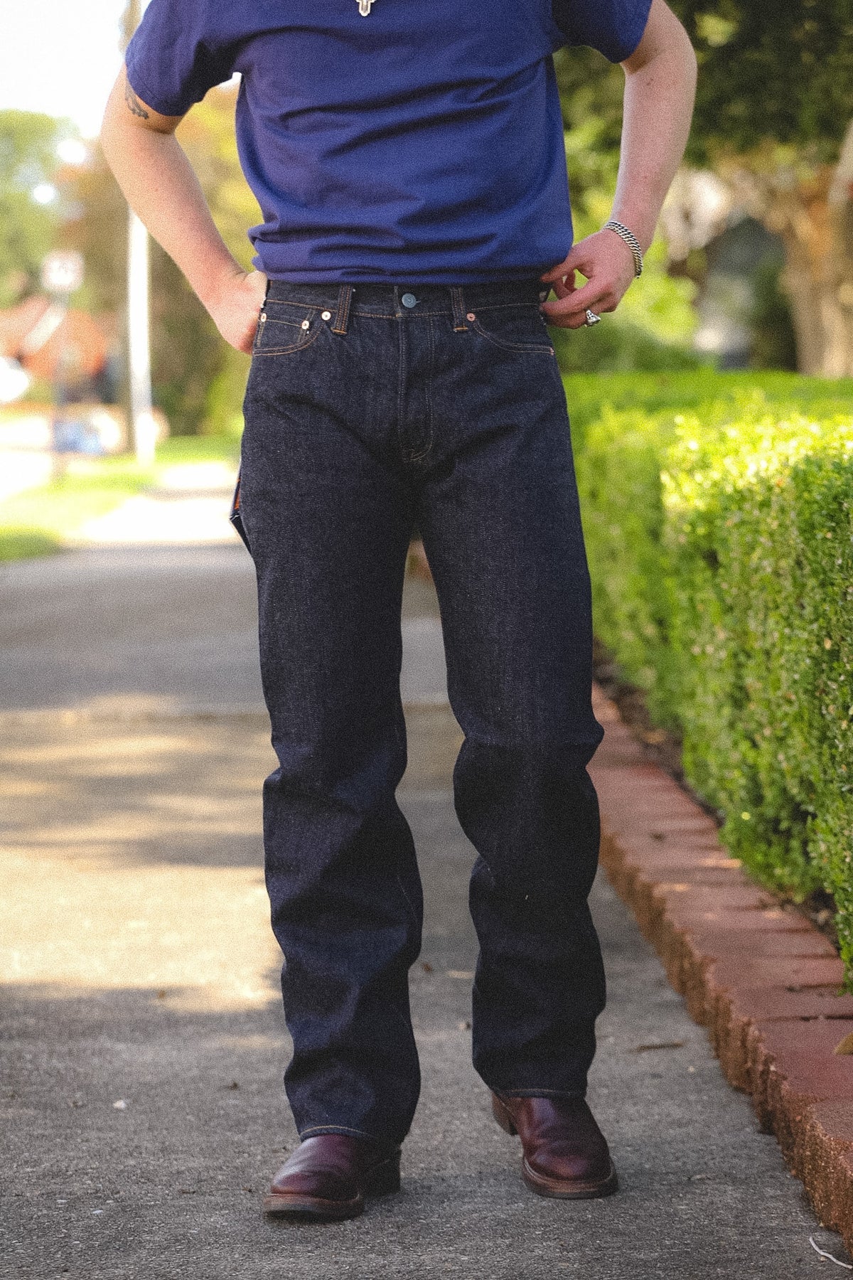 A person standing outdoors on a paved path wearing dark indigo denim jeans, a blue t-shirt, and brown leather shoes, with trimmed green hedges in the background.
