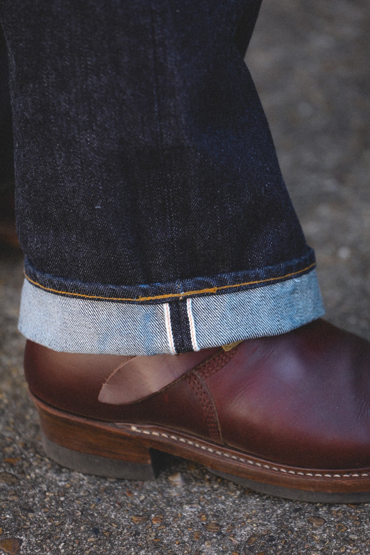 Close-up of a person's ankle wearing dark indigo jeans with a turned-up cuff showing a white stripe detail, paired with a brown leather shoe on a gray asphalt surface.