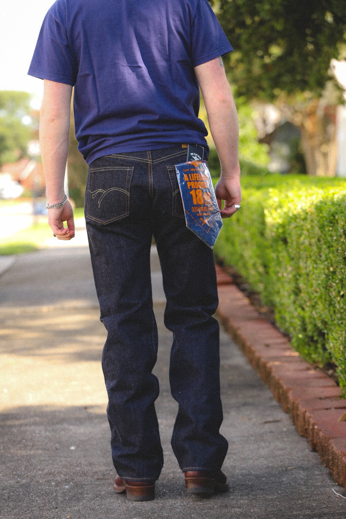 A person viewed from behind wearing dark blue denim jeans with a blue and orange paper tag attached to the back pocket, standing on a paved path outdoors next to a green hedge.