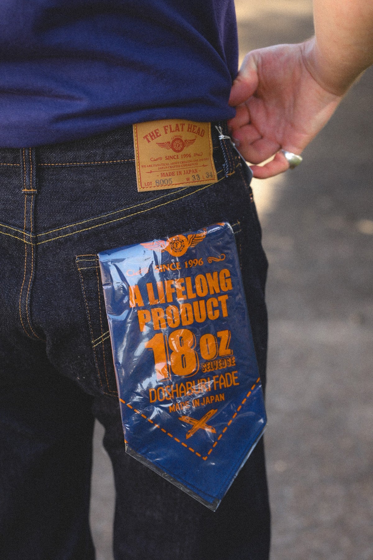 Close-up of a person wearing dark blue denim jeans, showing the back pocket with a large blue and orange novelty patch attached to the pocket, and part of the person's hand and torso visible.