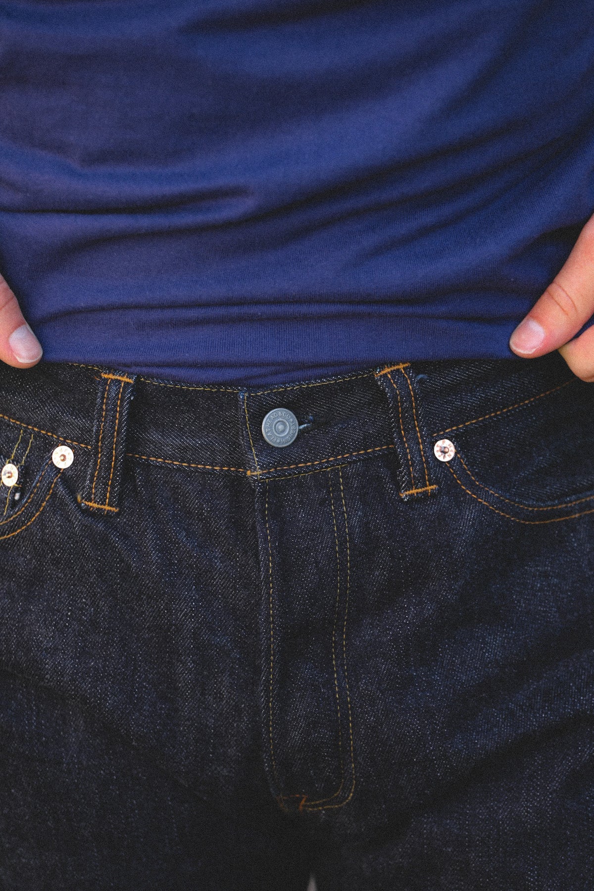 Close-up of a person wearing dark indigo denim jeans and a navy blue t-shirt, pulling the shirt up slightly to show the waistband and button of the jeans.