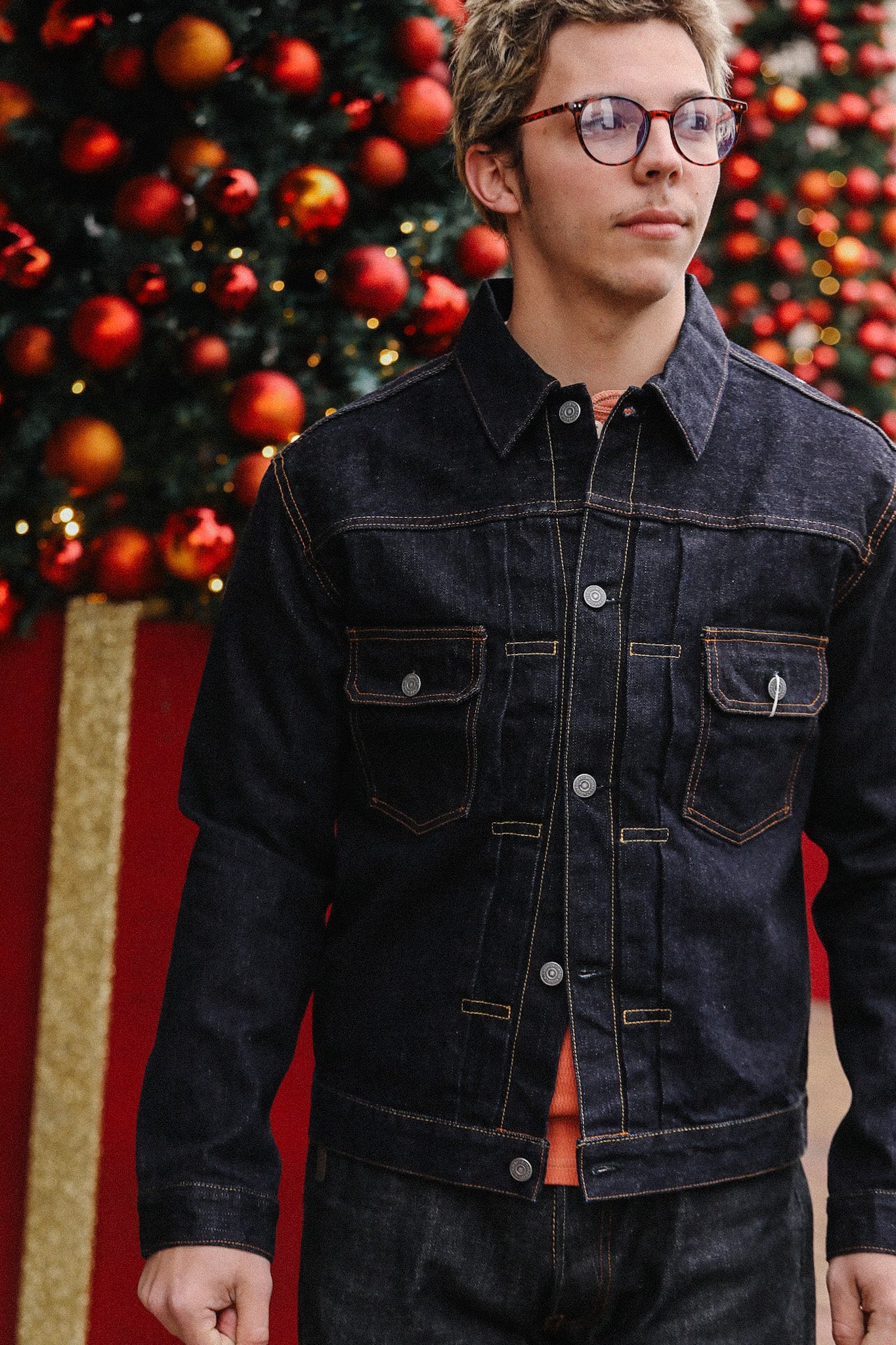 Man wearing black denim jacket with orange stitching in front of a Christmas tree decorated with red ornaments