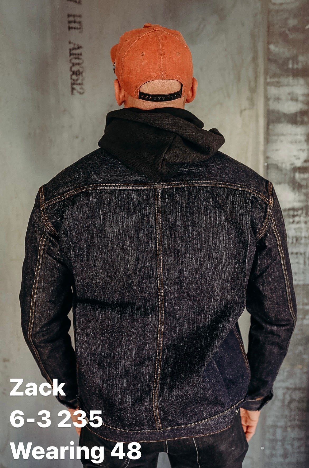 Man wearing dark denim jacket and black hoodie under rust-colored snapback cap, standing against concrete wall