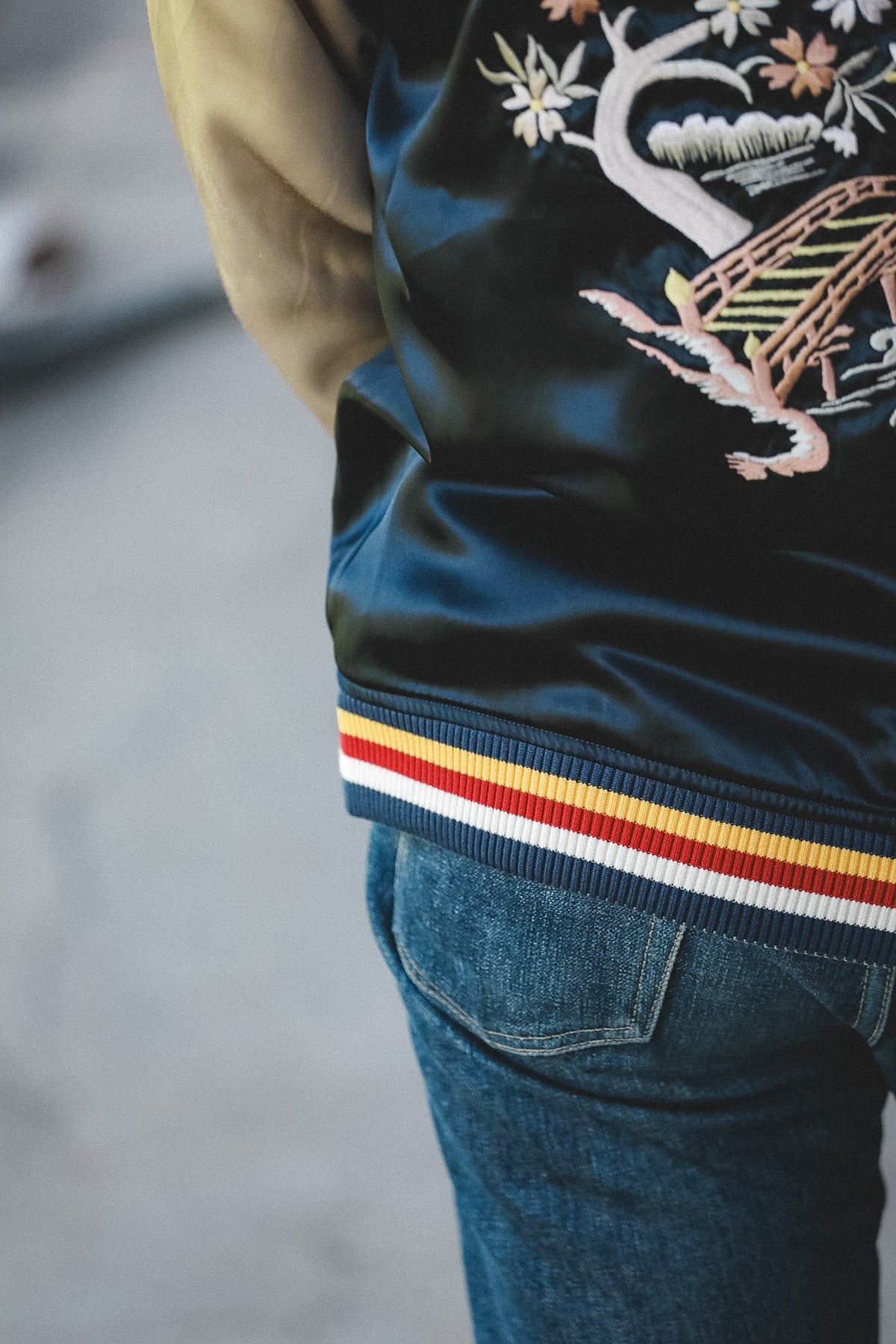 Close-up of a person wearing a dark navy satin bomber jacket with a tan sleeve, a multicolor embroidered dragon motif on the back, and a striped ribbed hemband in navy, yellow, and red, paired with blue jeans.