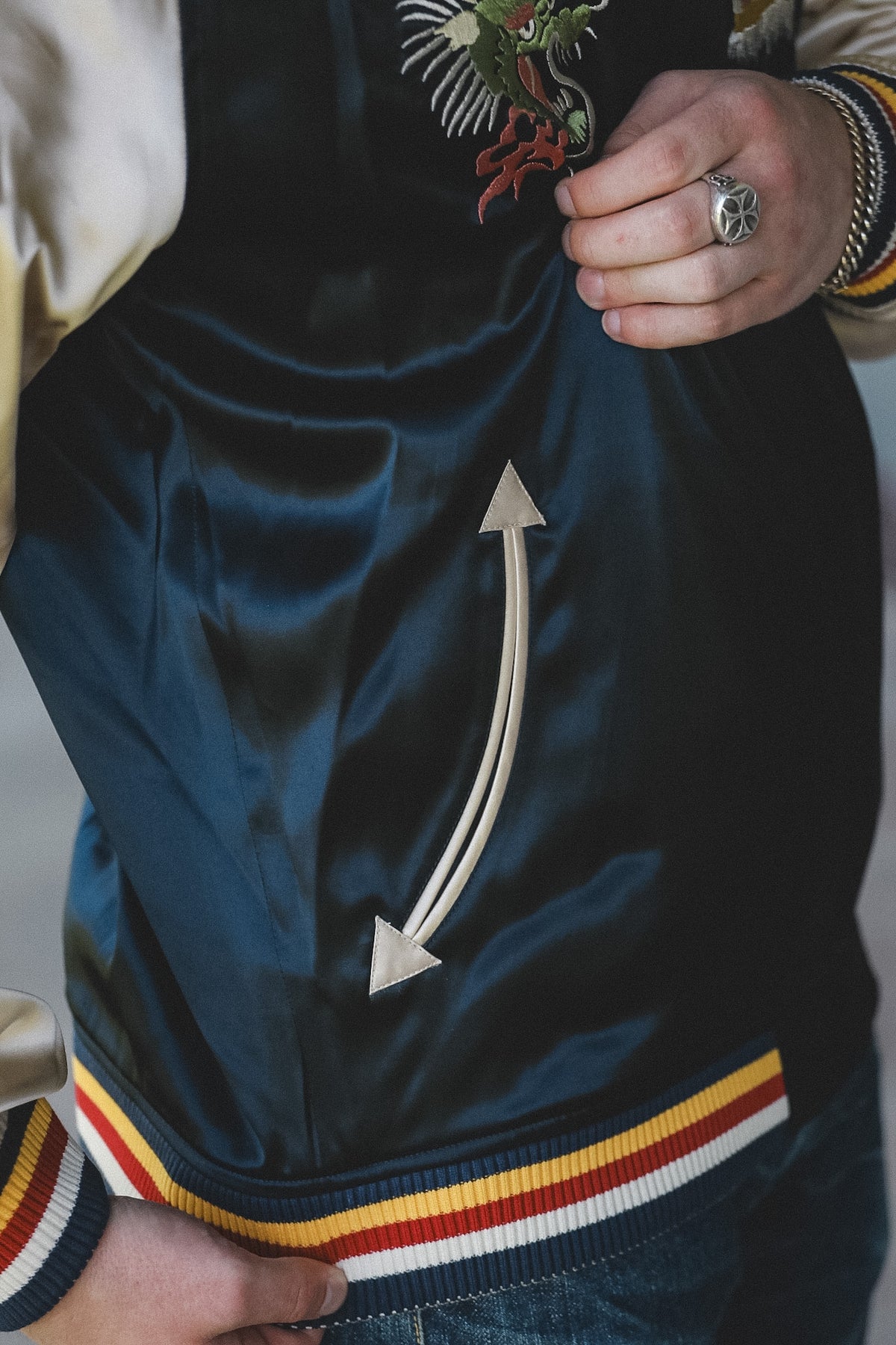 Close-up of a person wearing a dark navy satin bomber jacket with cream ribbed trim, a multicolor embroidered chest design, and a striped hemband with navy, cream, red, and yellow.