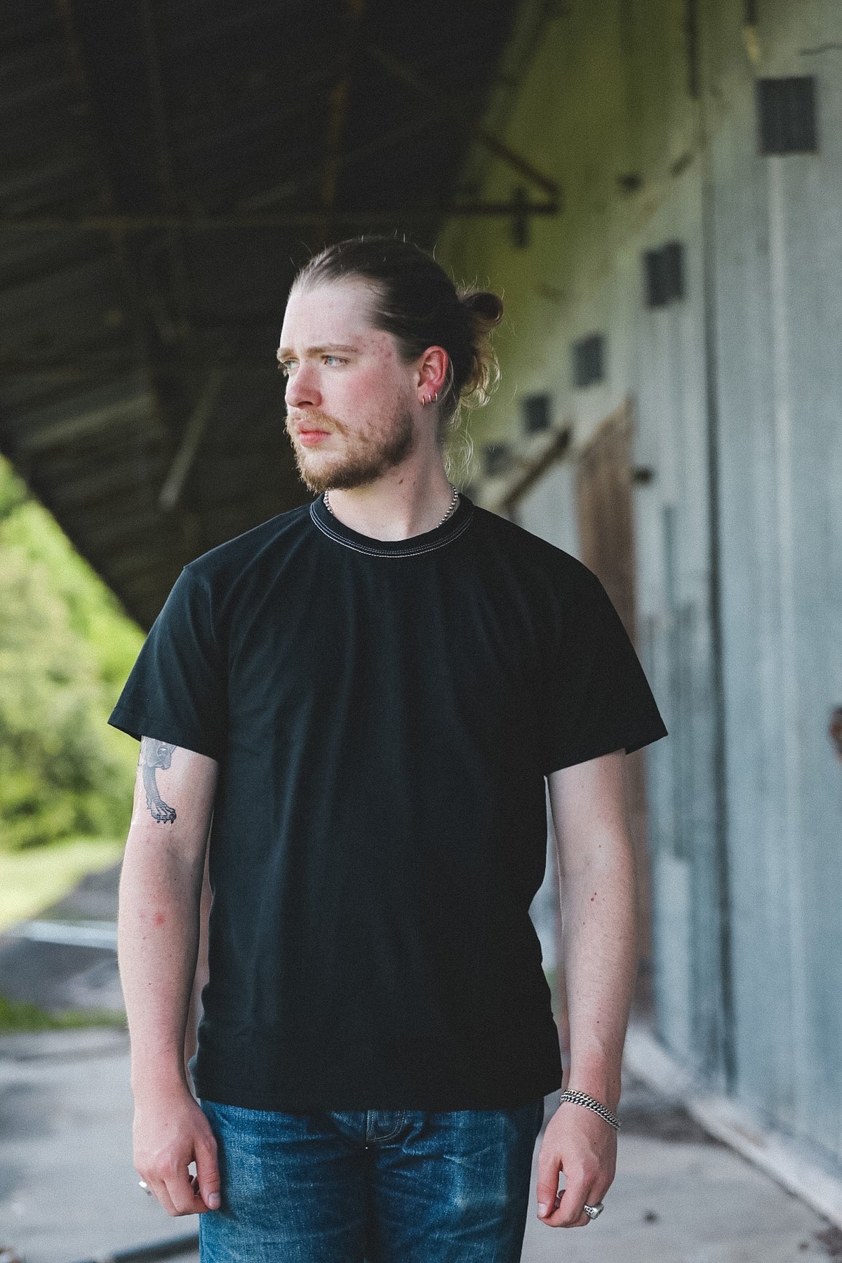 A man with a bun and beard wearing a black crewneck t-shirt and blue jeans, standing in a covered outdoor walkway next to a gray wall.